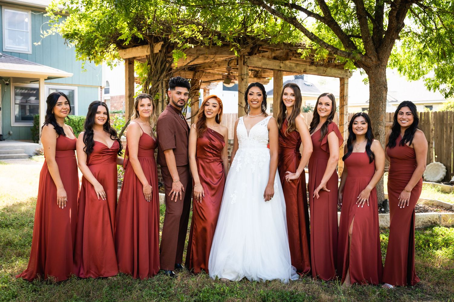 Wedding party poses outdoors wearing burgundy dresses with the bride in white.