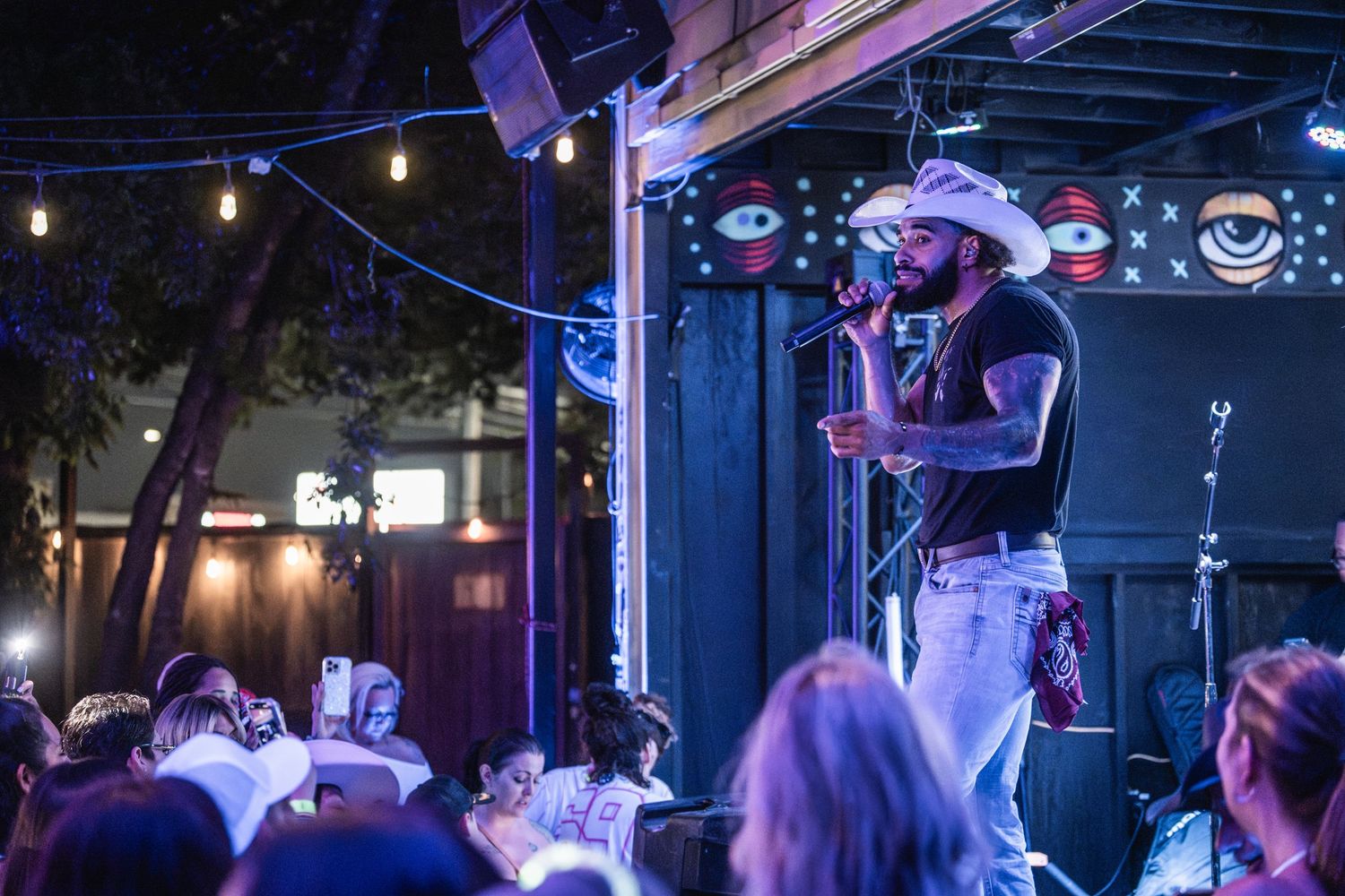 A performer wearing a white cowboy hat sings on stage at a nightclub with string lights and crowd in background.
