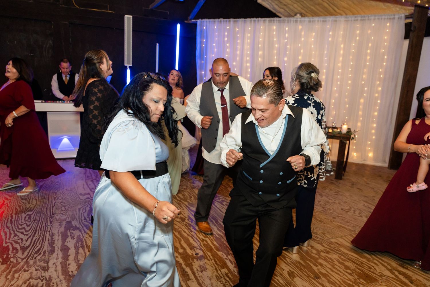 Energetic guests dancing together at a wedding reception with string lights and colored lighting in the background.