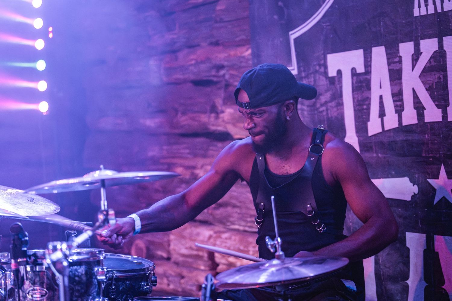Drummer performs energetically behind drum kit with purple stage lighting and rustic backdrop.