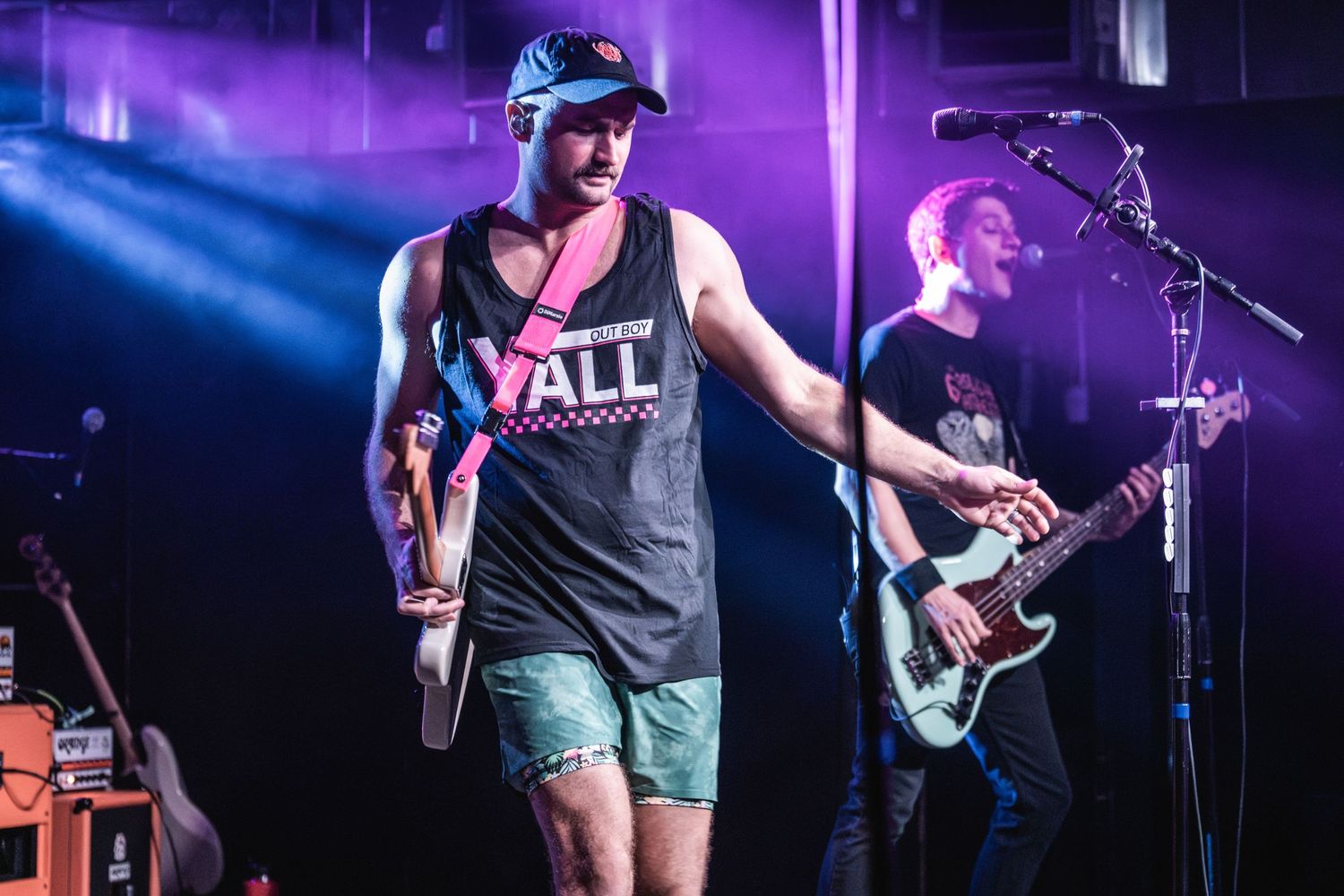 Energetic performer in a black tank top sings on stage with purple lighting and guitar player in background.