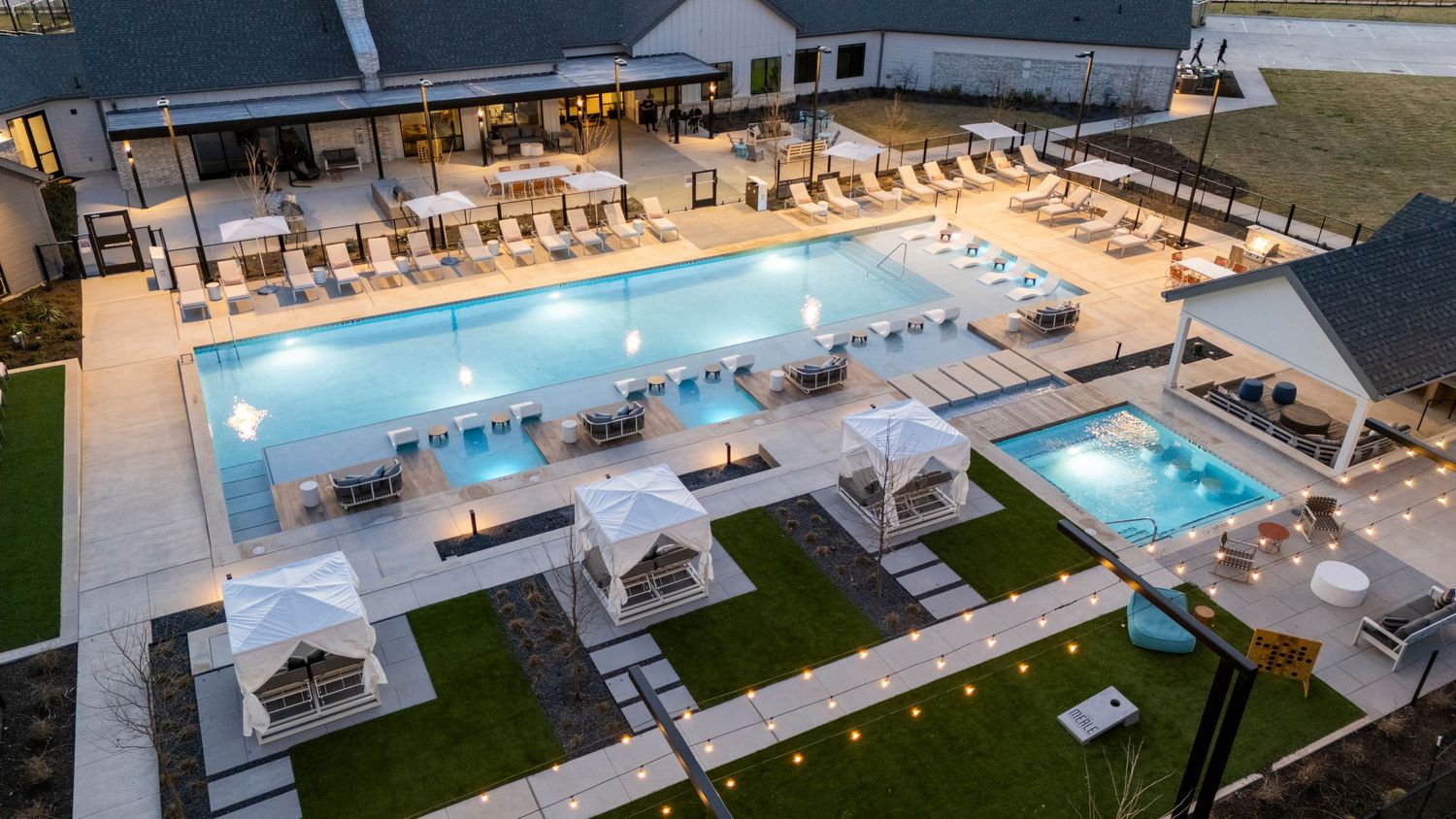 Aerial view of an illuminated luxury resort pool area with cabanas and lounge chairs at dusk.