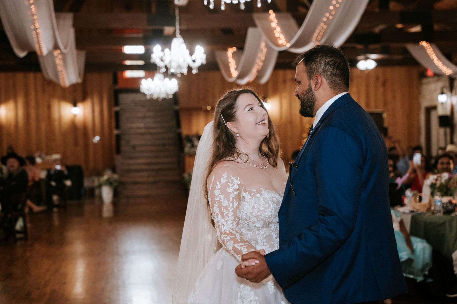 A couple shares their first dance in an elegant wood-paneled reception hall with crystal chandeliers and draped decor.