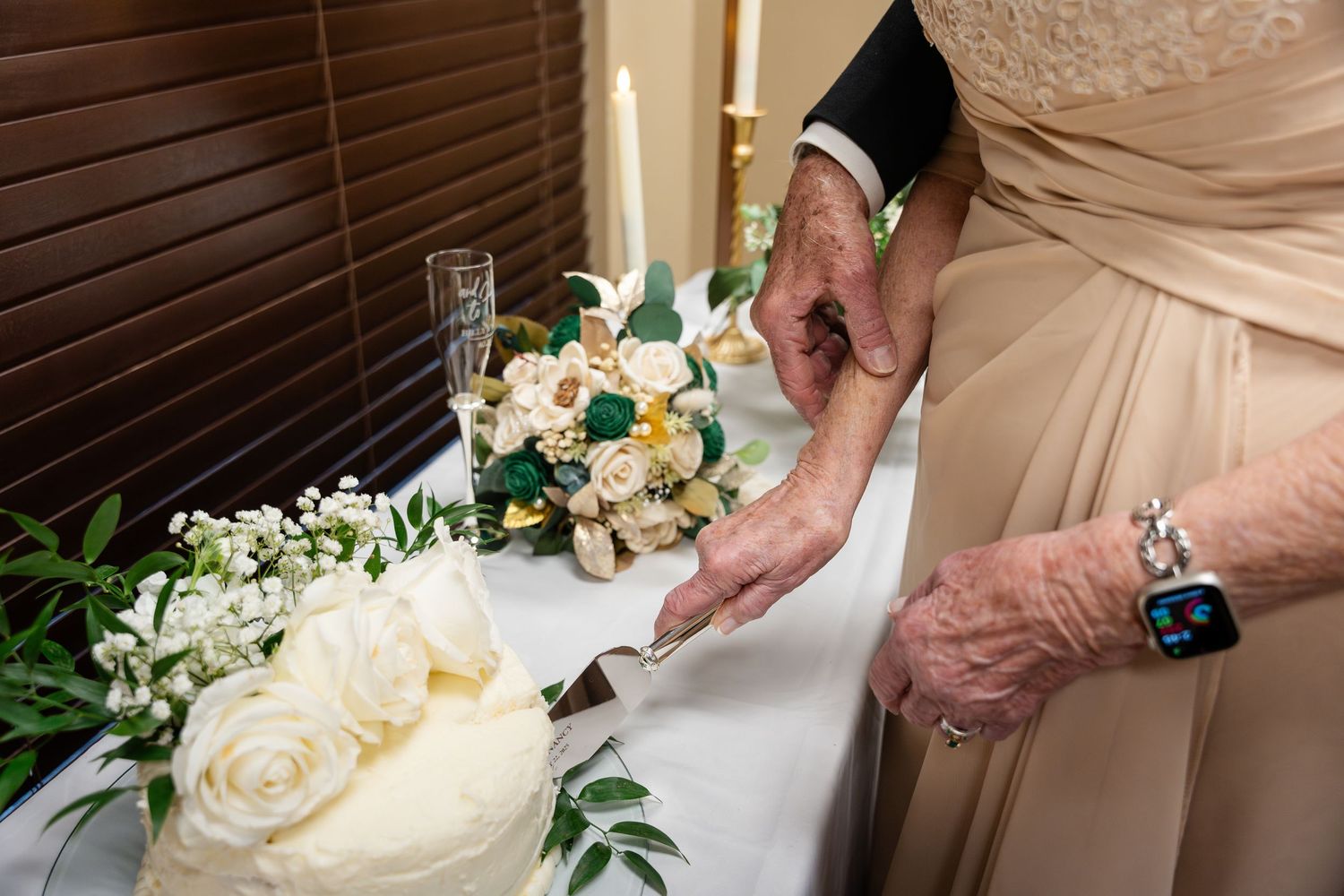 Hands cut into an elegant white wedding cake beside a bouquet of white and peach roses with greenery.
