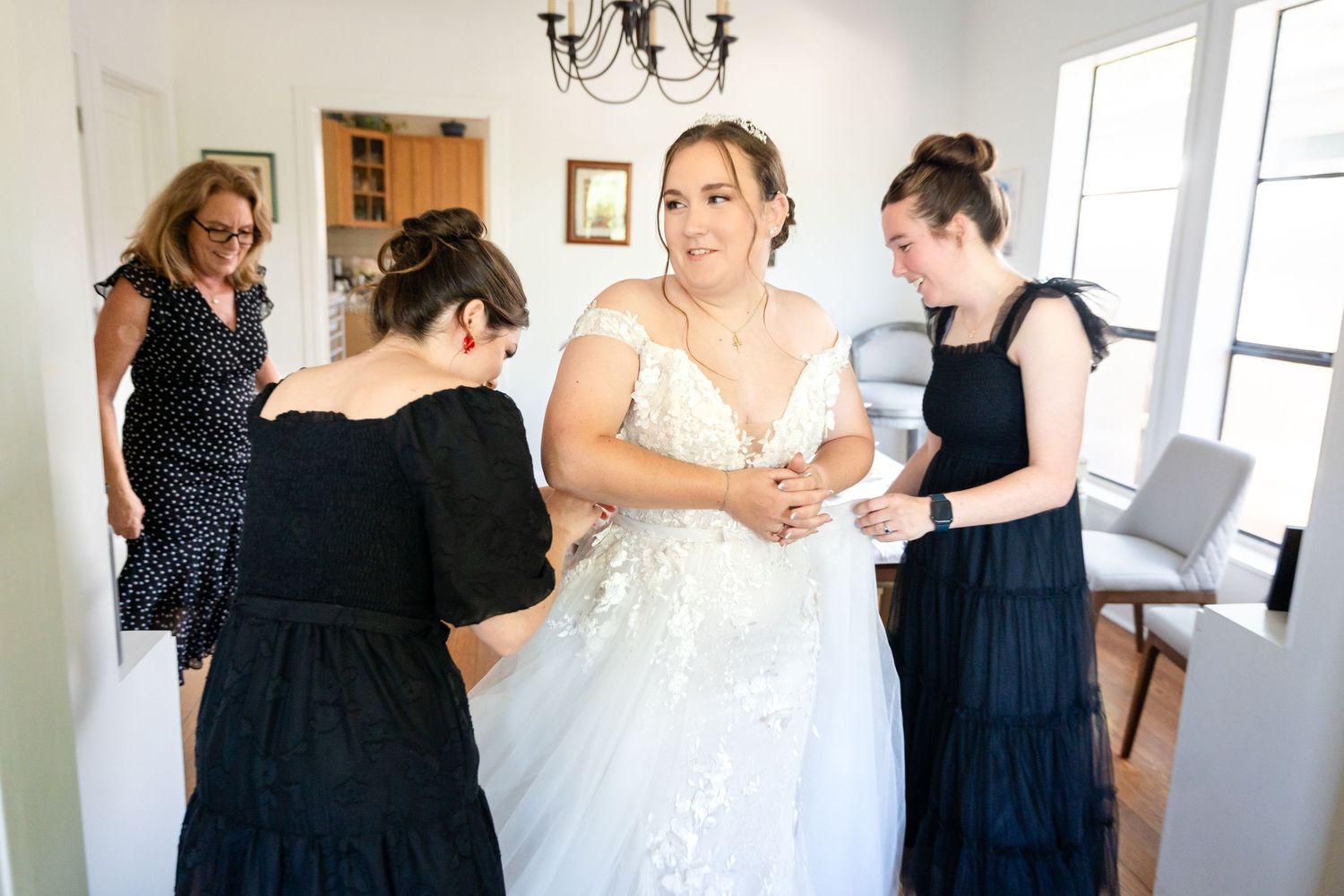 Wedding party members assist with final dress adjustments in a bright indoor space.