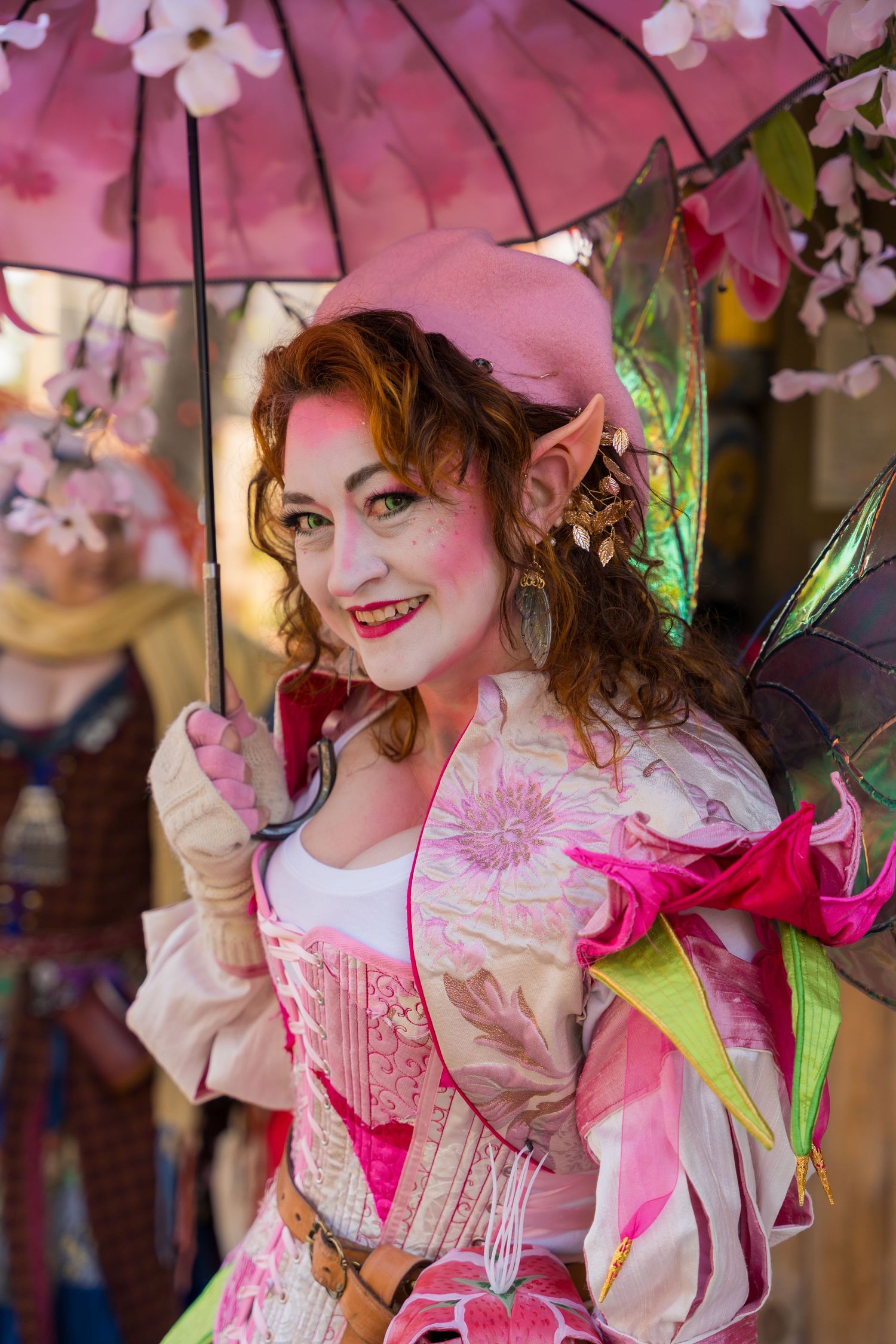 Fairy character with pink parasol and wings poses in floral costume at renaissance festival.