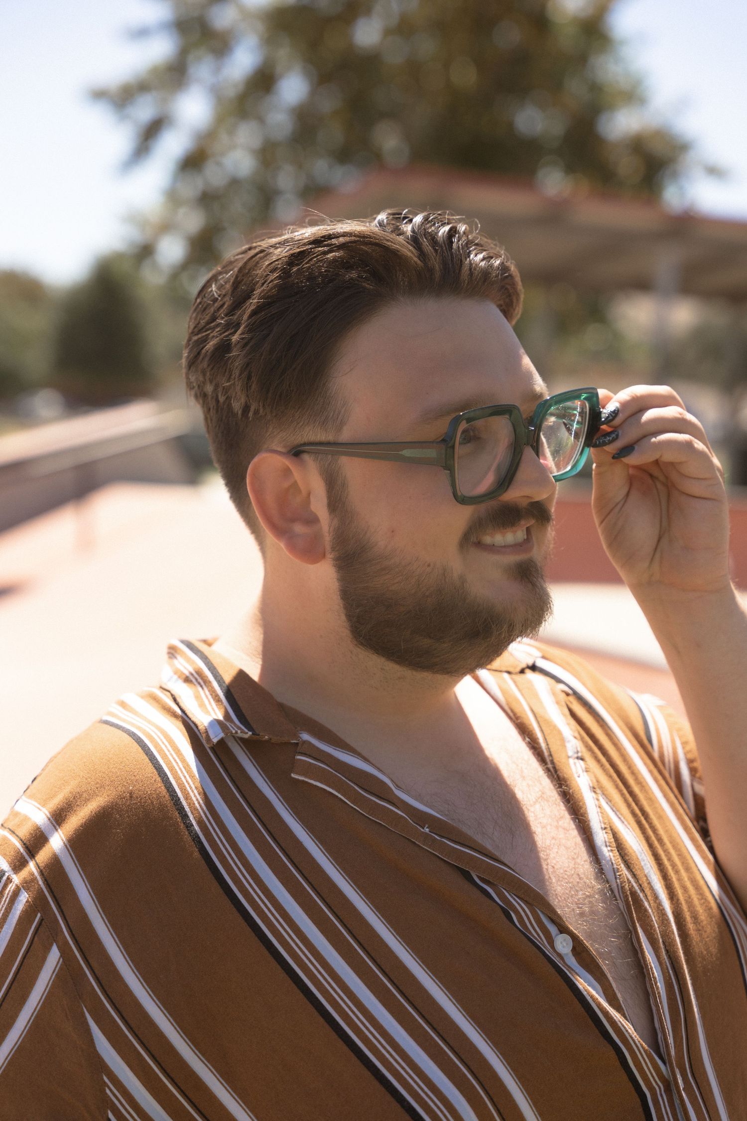 A person in glasses and a striped shirt smiles while adjusting their eyewear in warm sunlight.