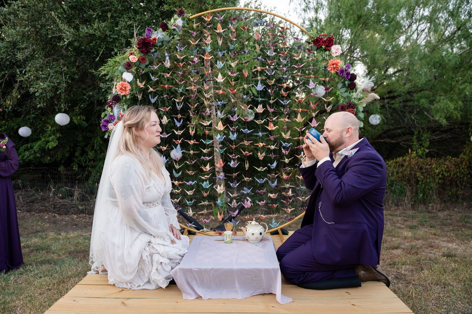 A couple shares a traditional tea ceremony under a decorative arch with hanging paper butterflies.