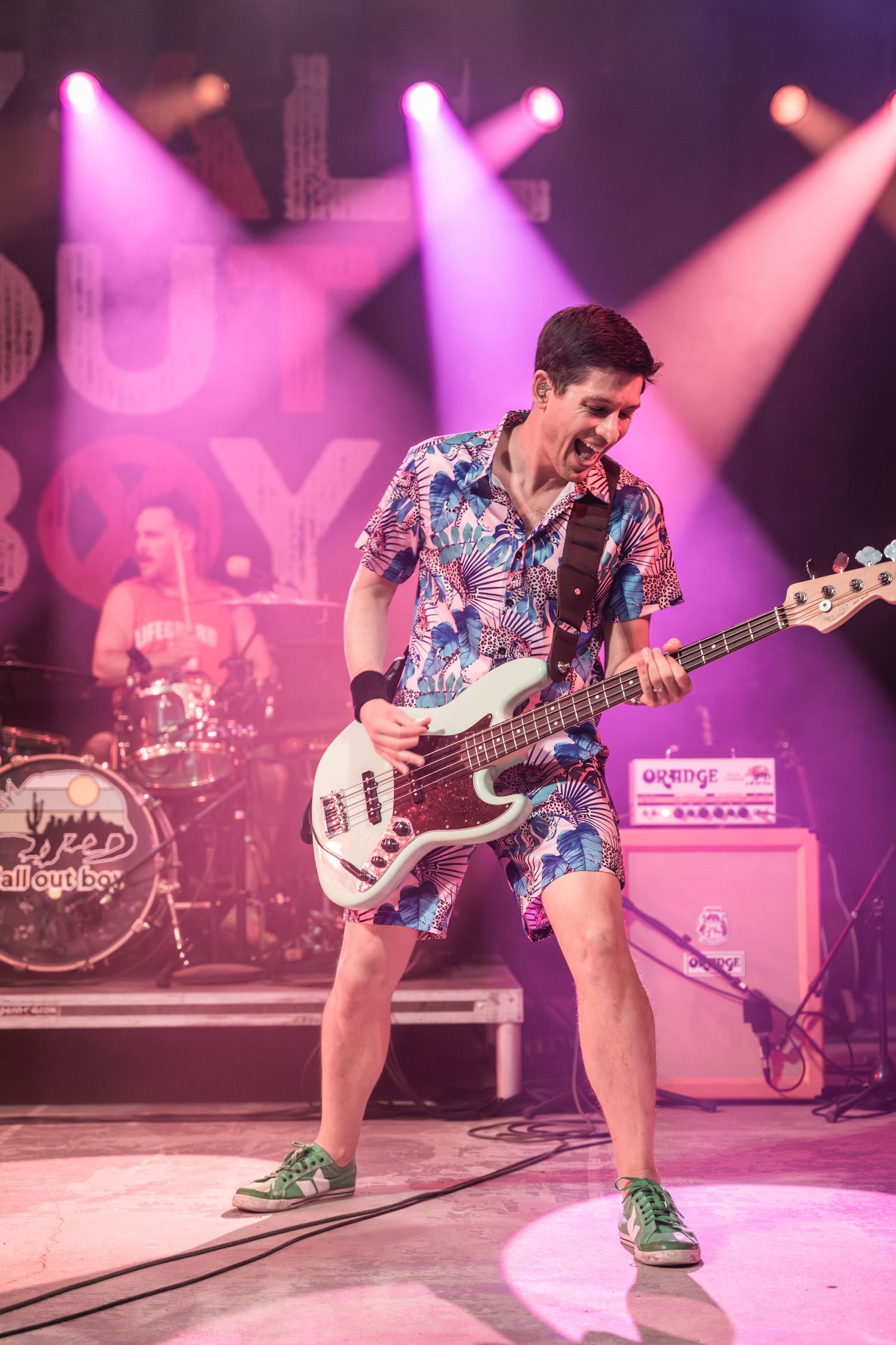 Bass player in floral shirt and shorts performs energetically under purple stage lights.