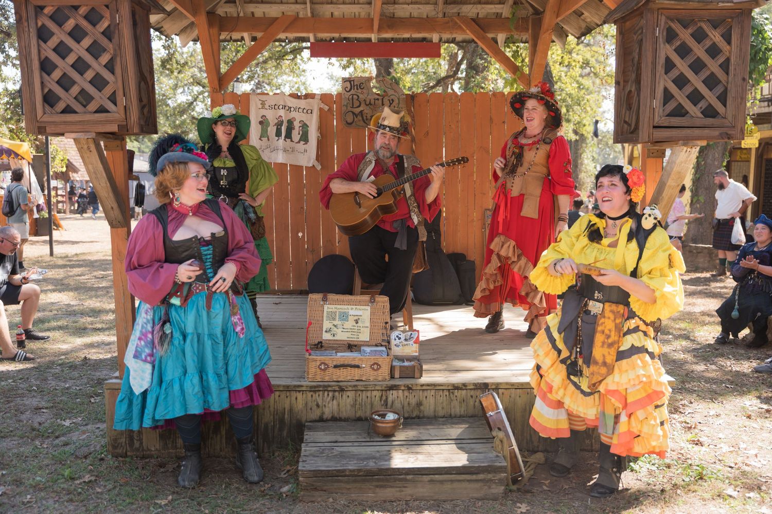 Performers in colorful renaissance costumes entertain guests at a wooden gazebo during a fair.