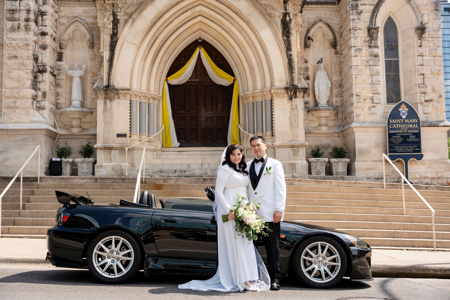 Two people stand next to a sleek black sports car in front of a beautiful stone church with gothic architecture.