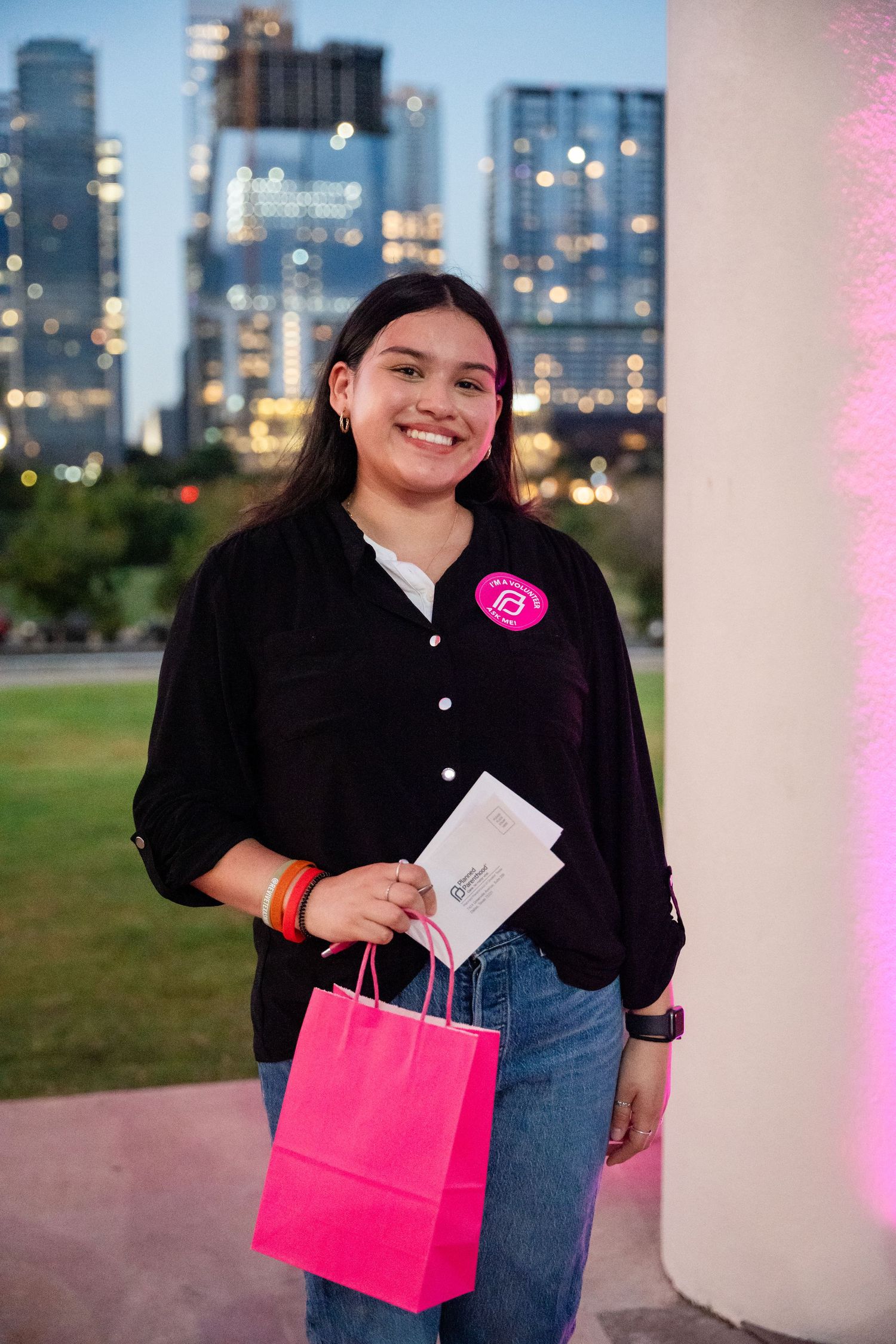 An attendee poses with a pink gift bag at an outdoor evening event with city buildings in background.