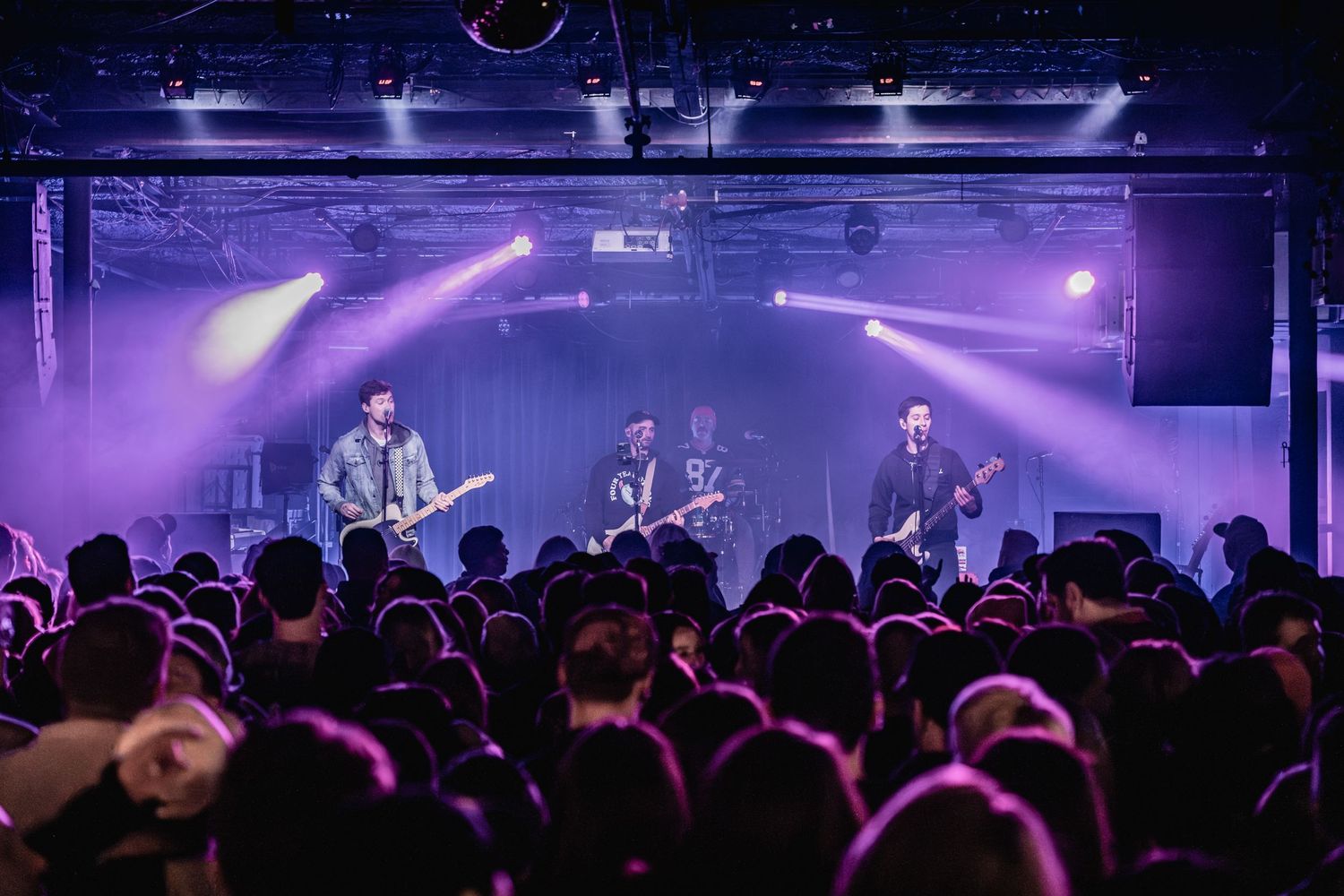 Band performs on stage with purple lighting effects as crowd watches at indoor concert venue.