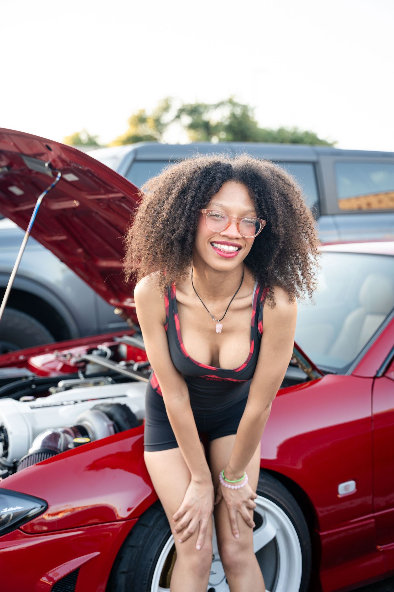 Model with curly hair poses cheerfully next to a red car with open hood at auto show.