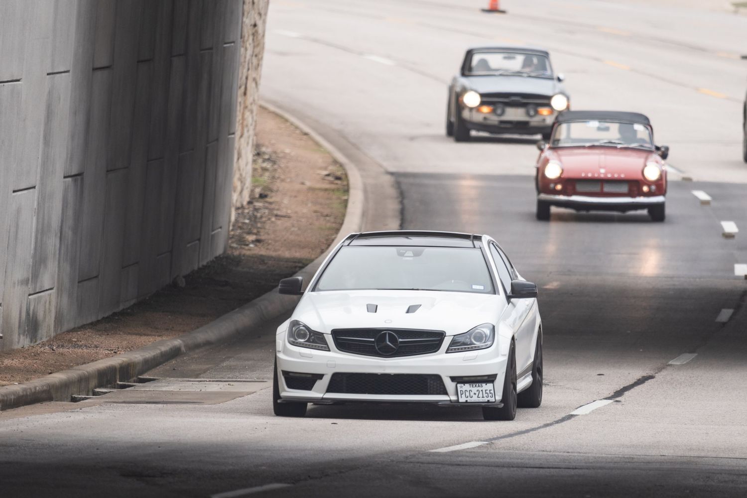 White sports car leads through an underpass followed by vintage cars on a track.