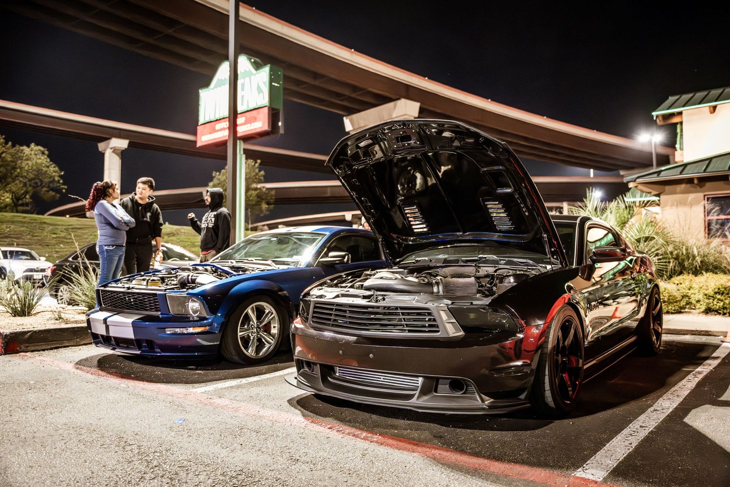 Two modified sports cars with open hoods parked under covered parking at night.