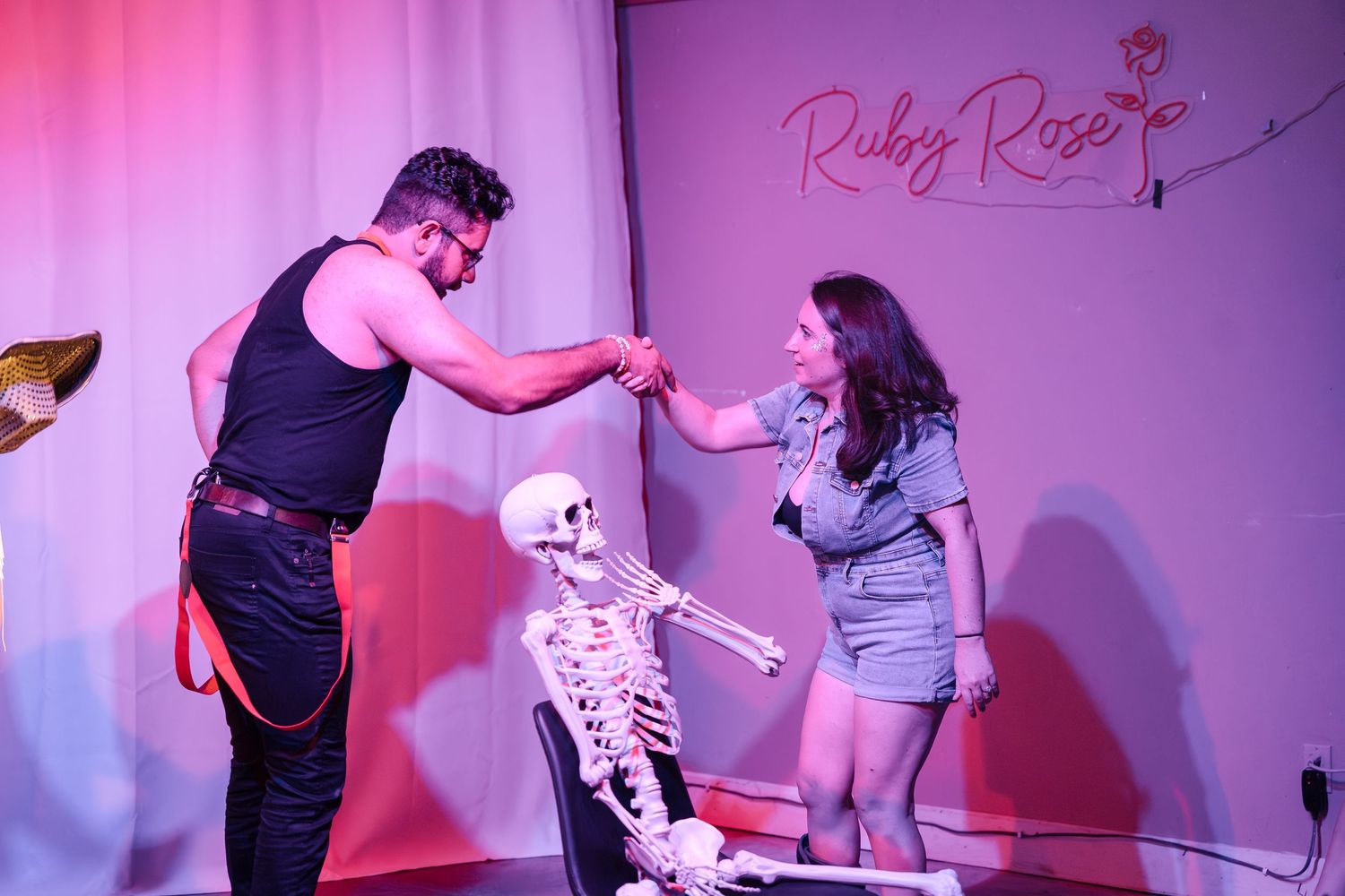 Performers interact with skeleton prop on pink-lit stage.
