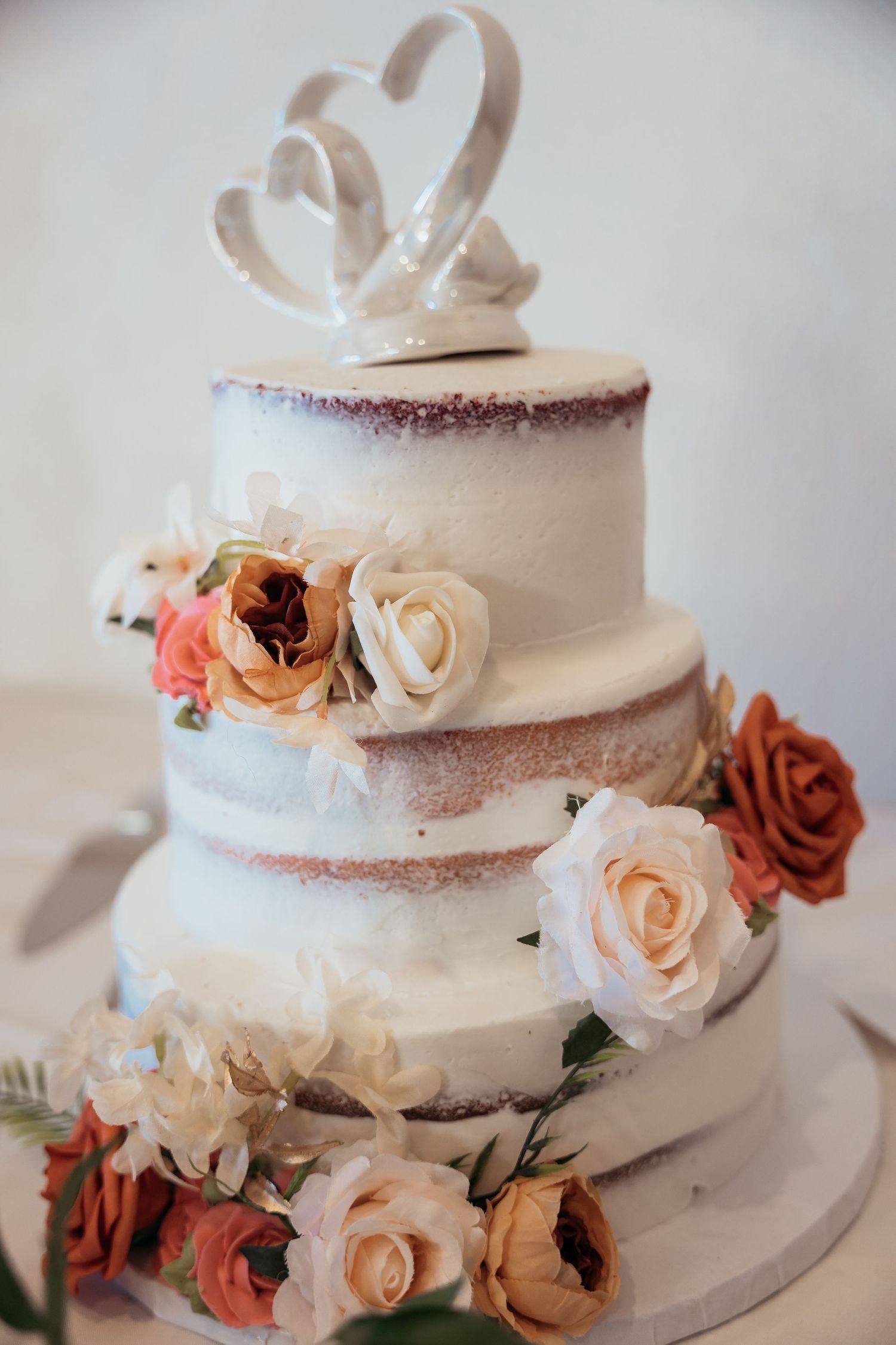 Three-tiered white wedding cake decorated with pink and peach roses and a heart-shaped topper.