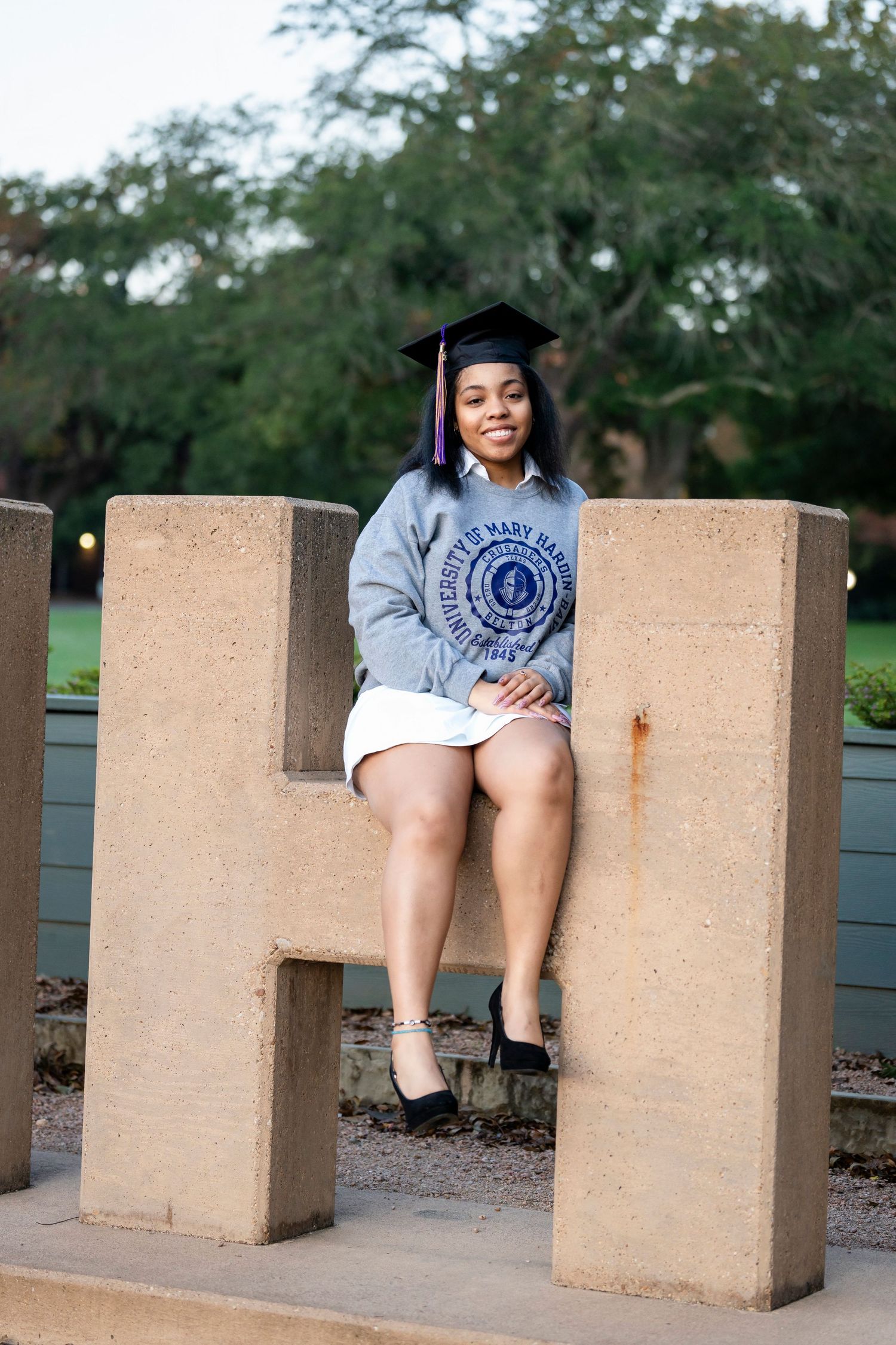 Graduate in academic regalia sits on large concrete letter H during outdoor photo session.
