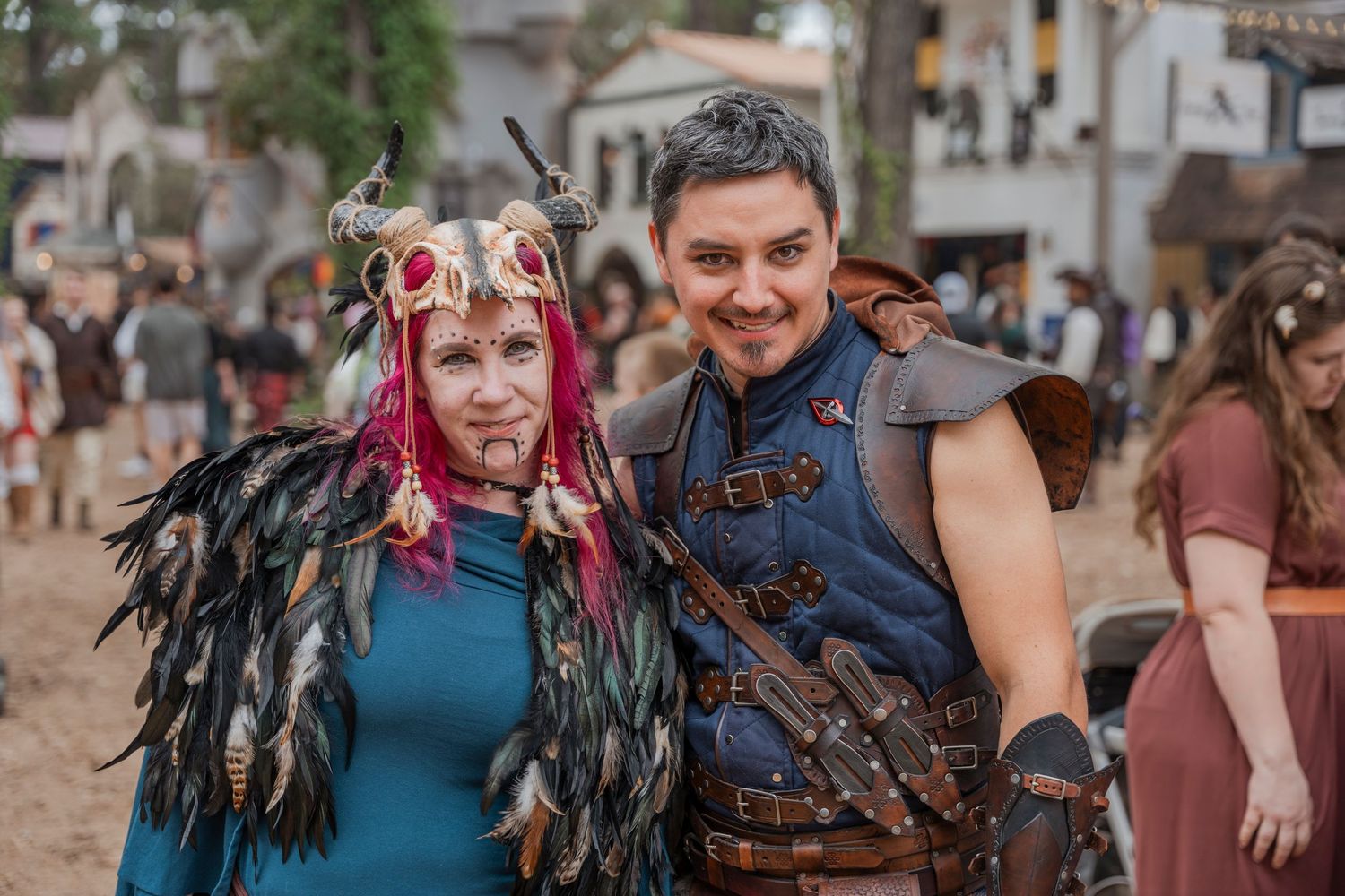 Two festival attendees in medieval fantasy costumes with feathers and leather armor at a renaissance faire.