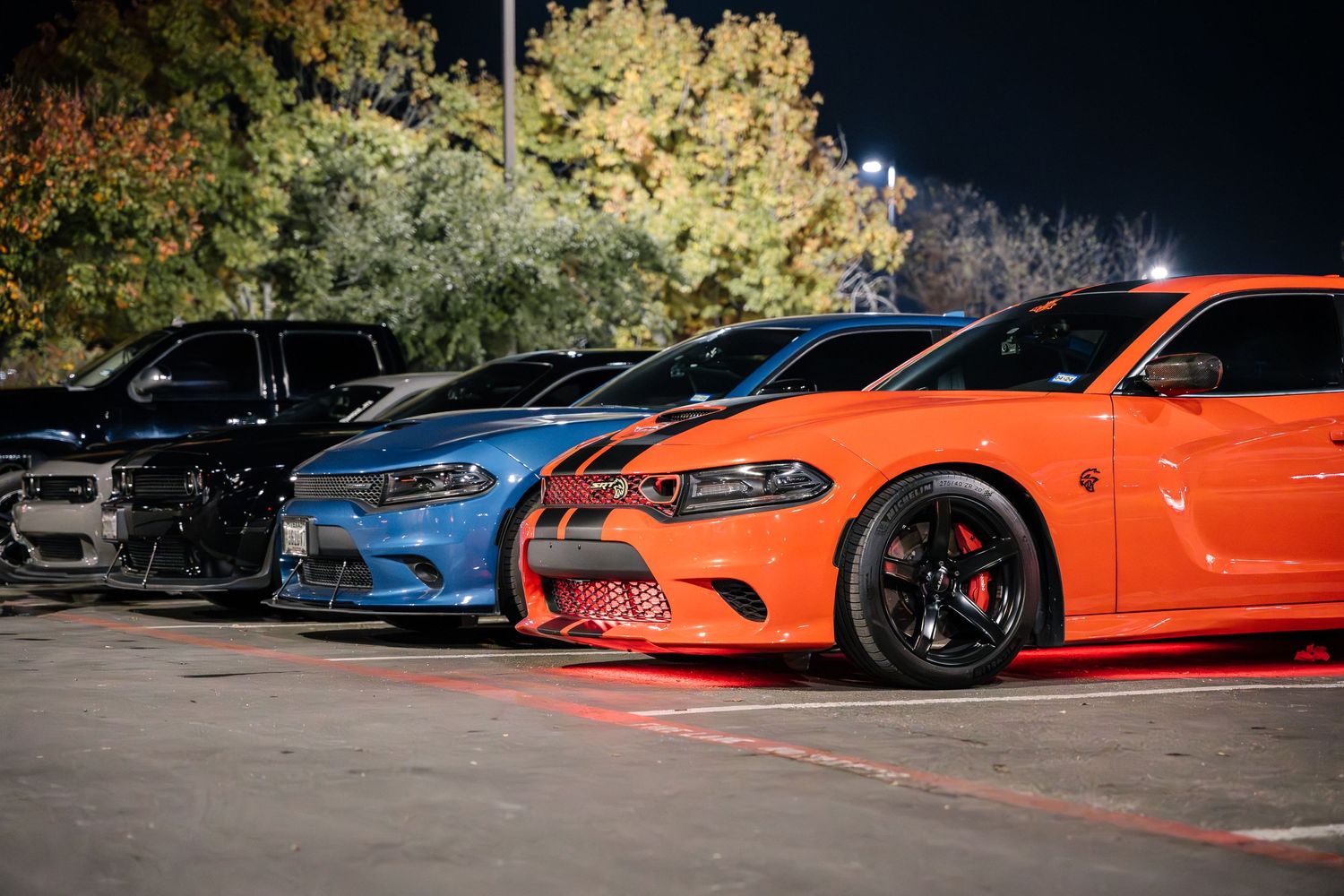 Modern Dodge muscle cars in blue and orange showcase custom modifications in a nighttime parking lot.