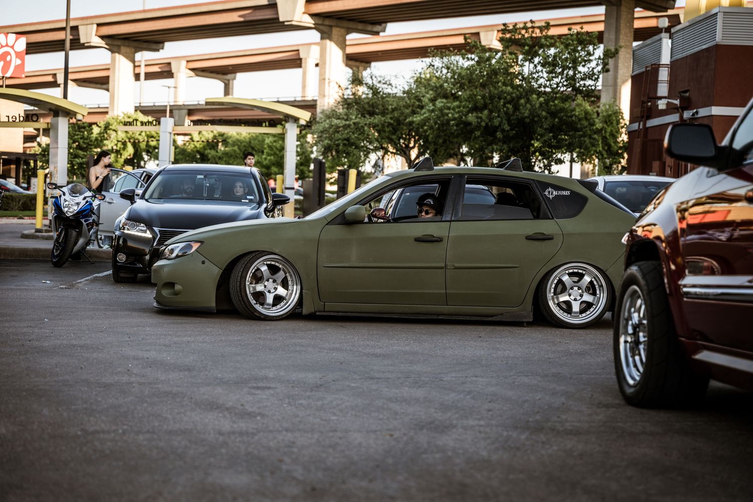 Olive green modified sedan with lowered suspension and custom wheels in parking lot.