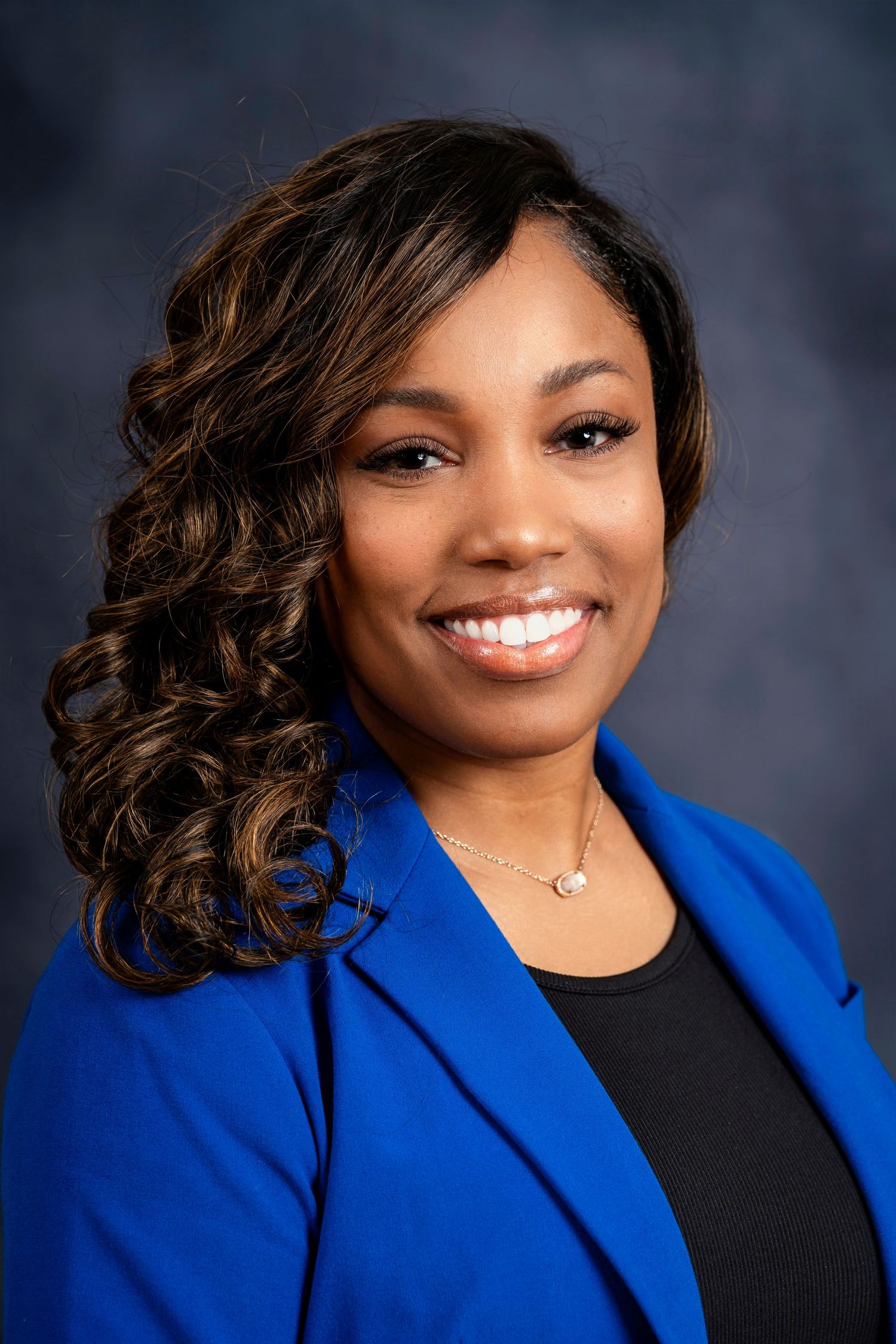 Professional portrait of a person in bright blue blazer with curled hair against dark background.