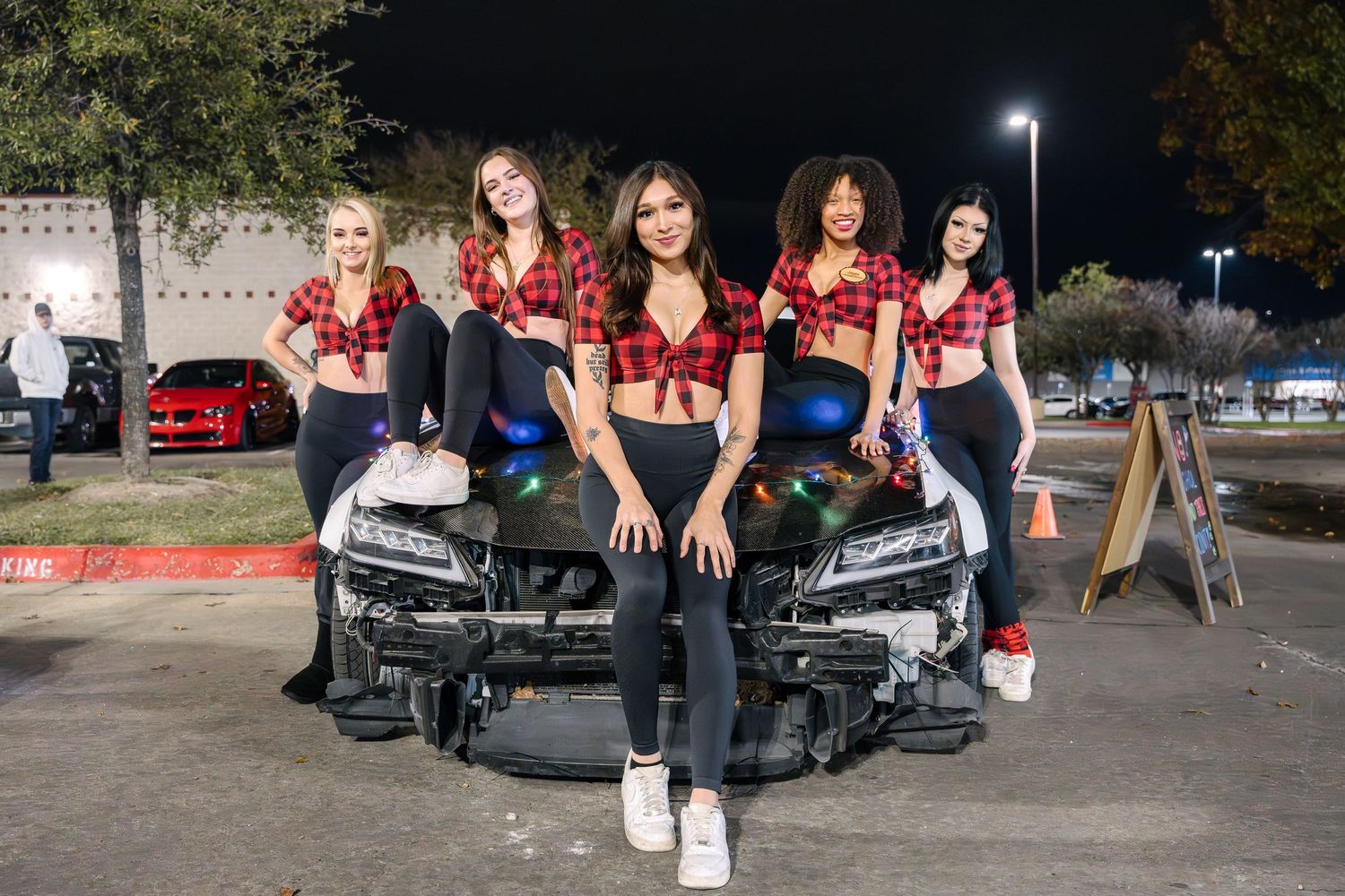 Women in matching red crop tops pose on a black car at night.