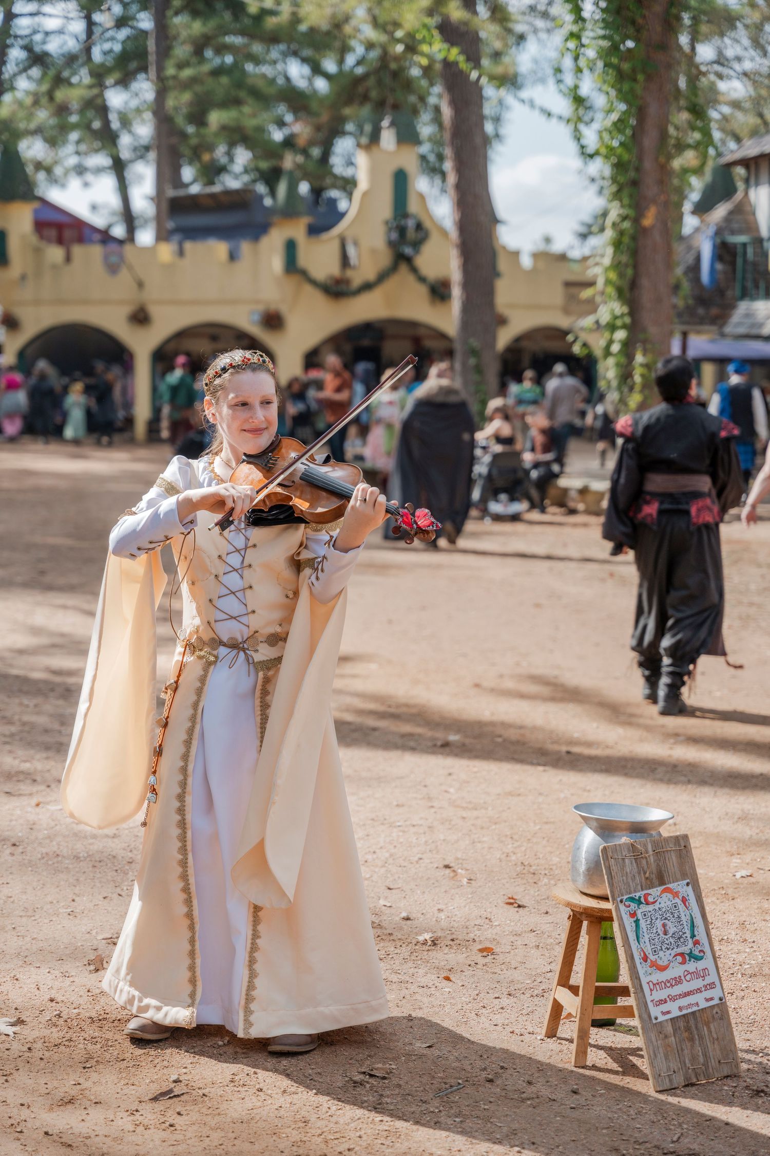 Musician in elegant white renaissance costume plays violin on dirt path at medieval festival.