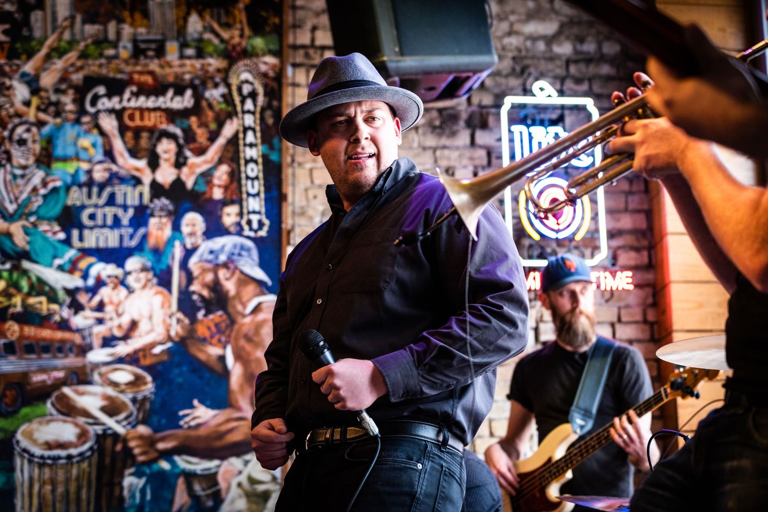 Performer in black attire and hat entertaining crowd at intimate brick-walled venue with colorful backdrop.