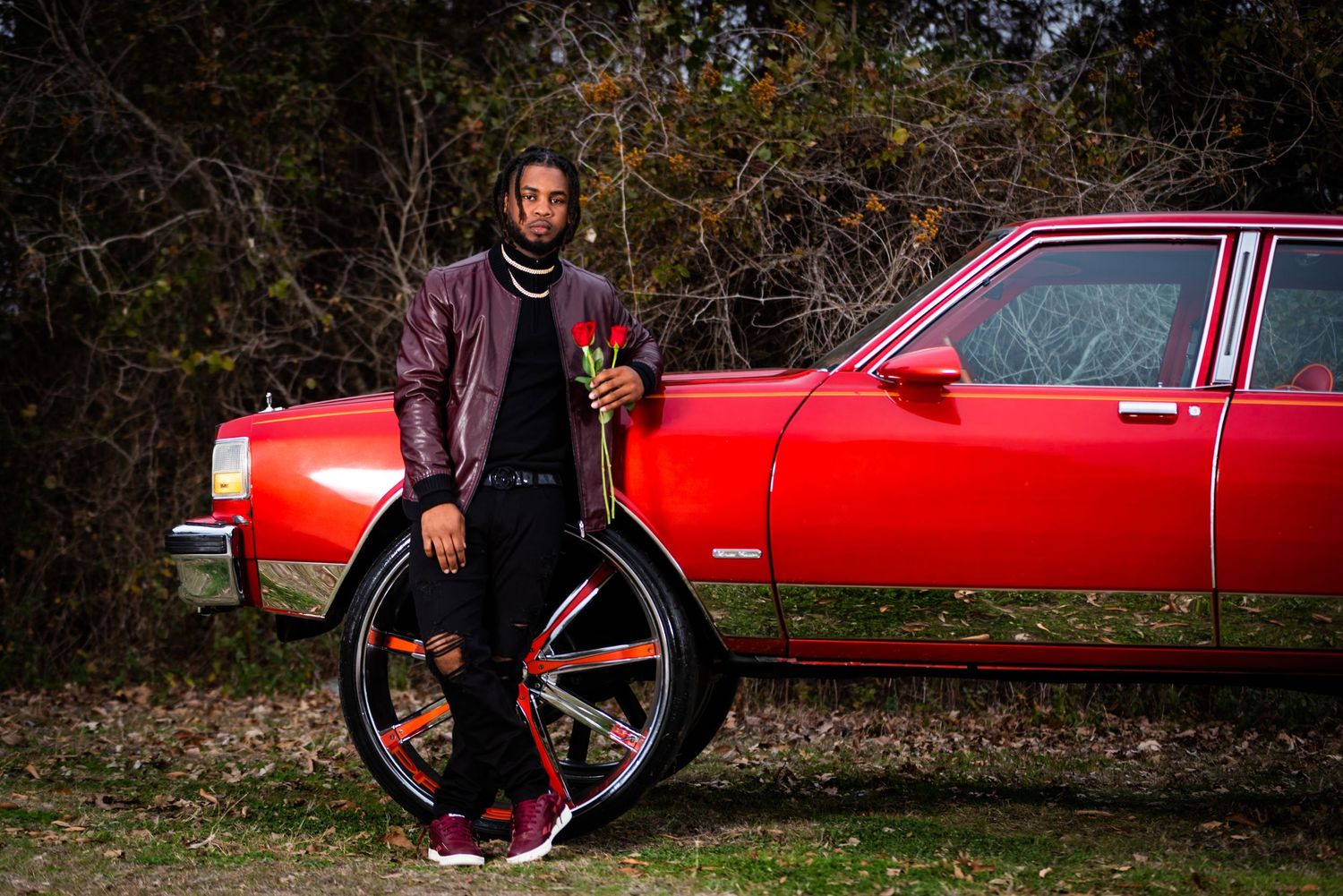 A person poses with a vintage red car and large custom wheel in an outdoor setting.