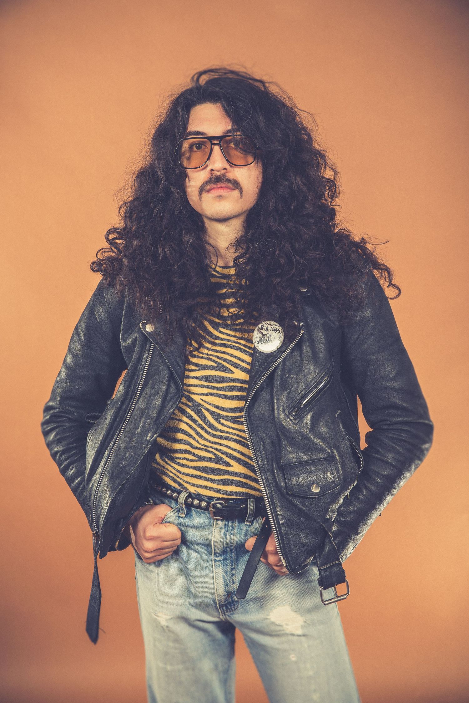 Retro-styled portrait with leather jacket, tiger print shirt and long curly hair against orange backdrop.