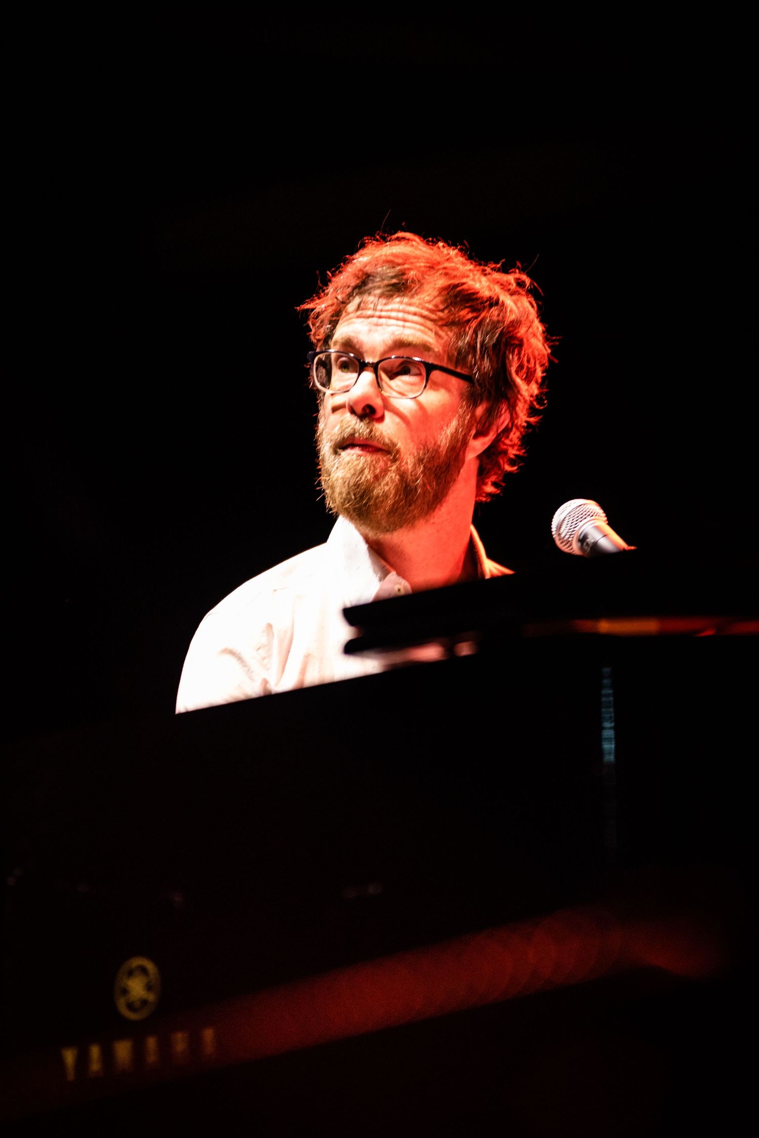Pianist with beard and glasses performs under dramatic red lighting.