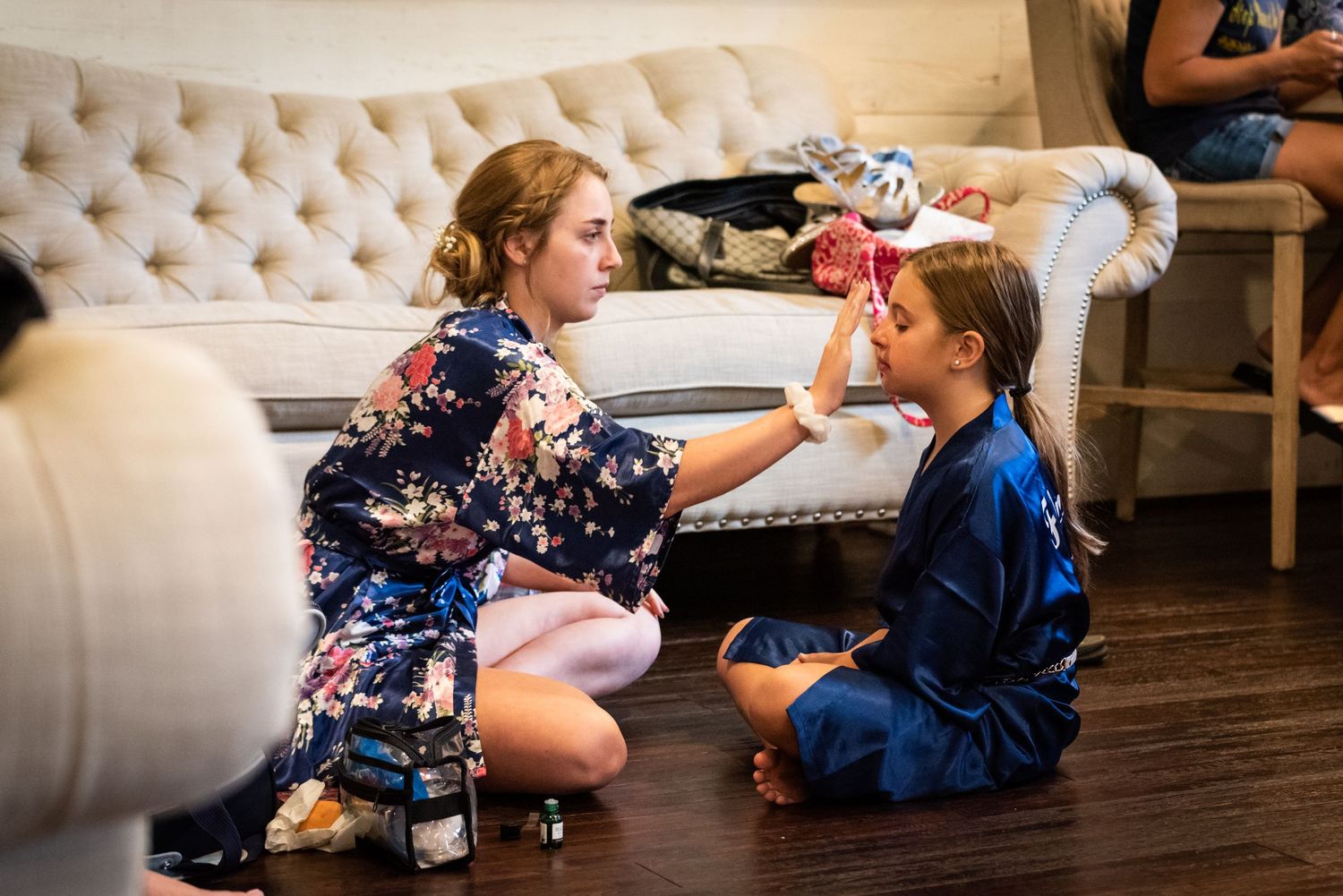 Two people sit on the floor, one applying makeup to the other.