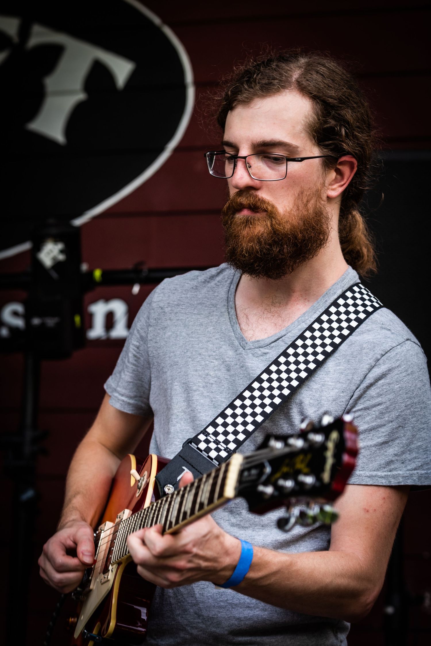 Musician playing guitar with checkered strap against red wall backdrop.