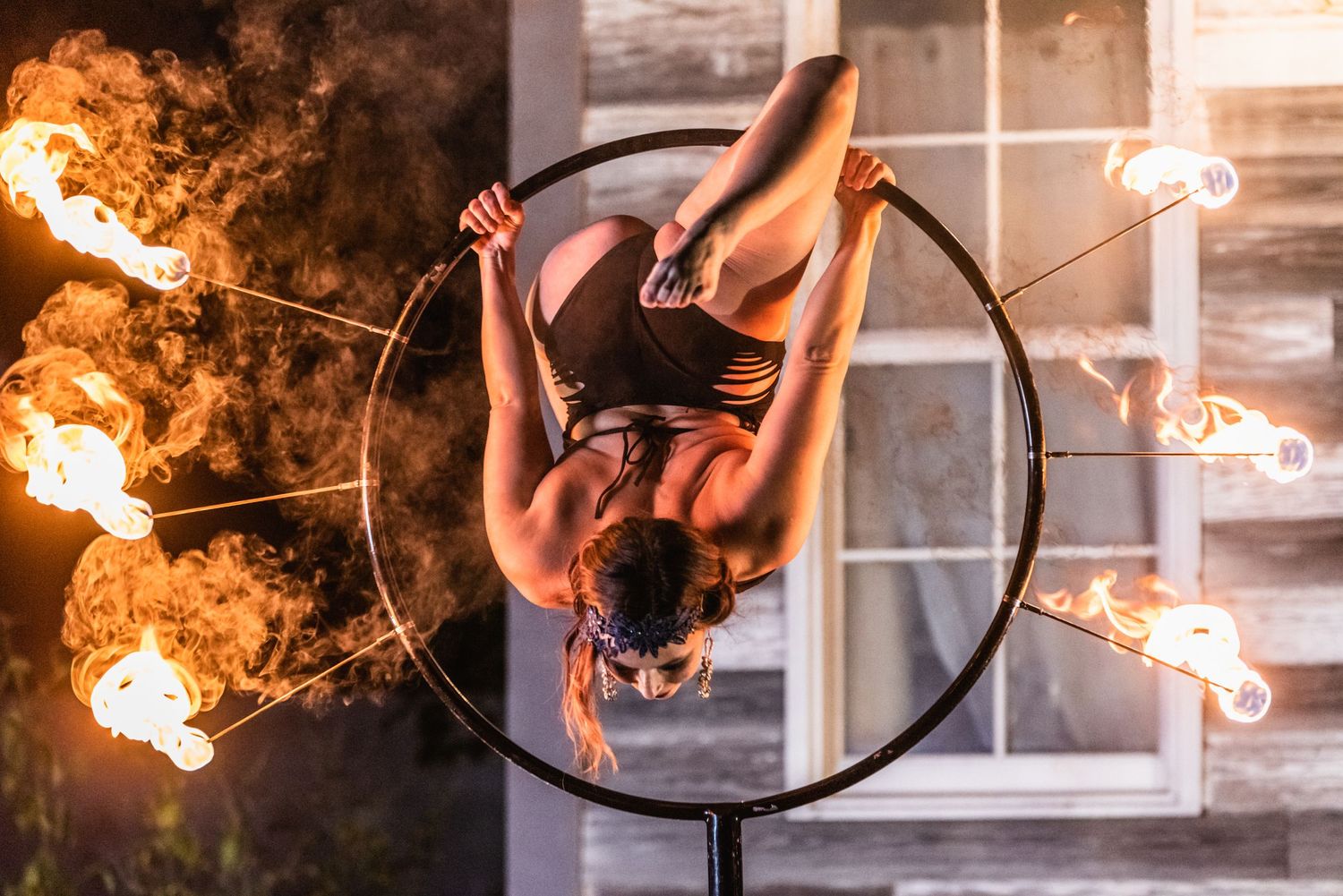 Aerial acrobat performs inverted position on hoop surrounded by fire torches at night.