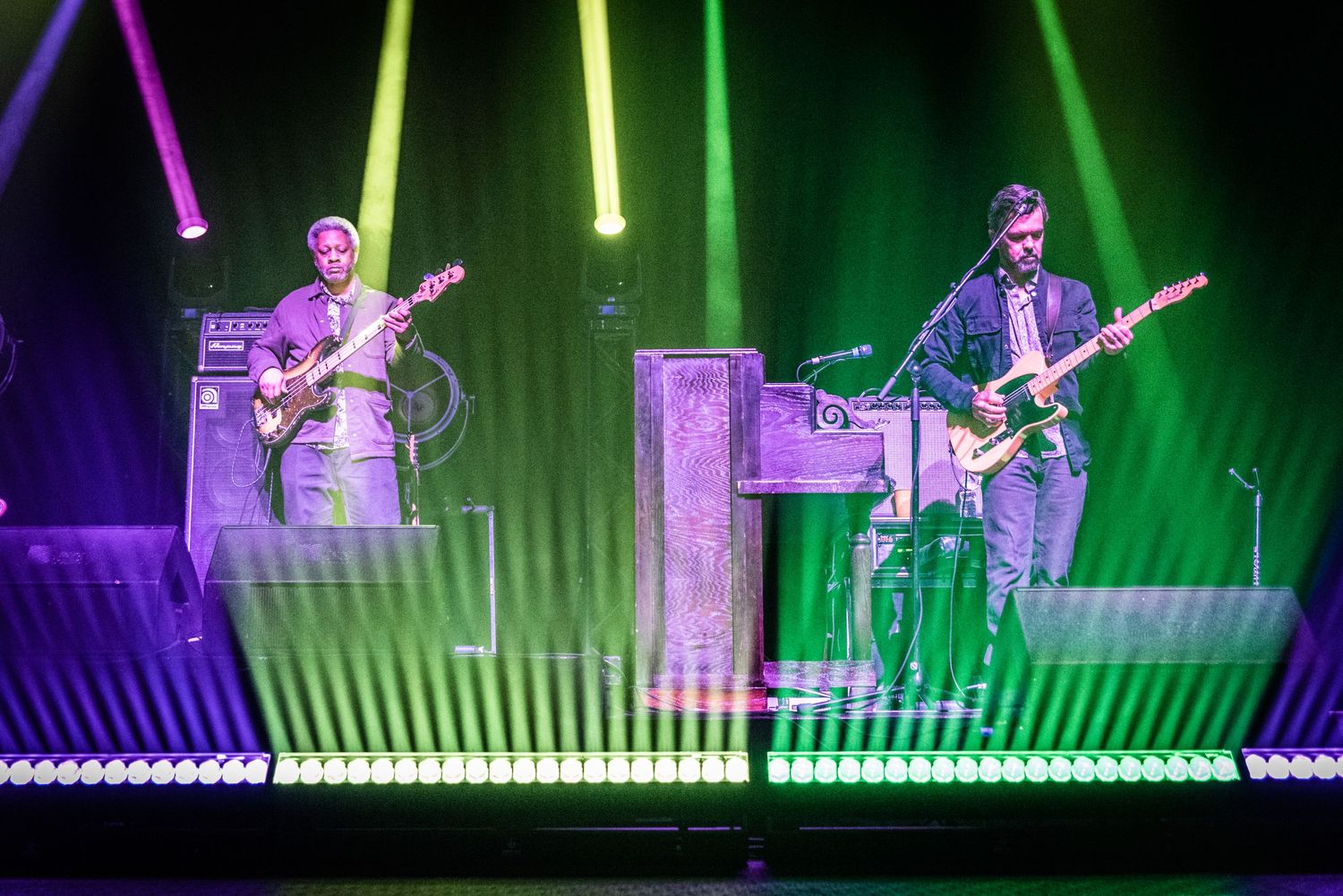 Musicians performing on stage with vibrant purple and green light beams creating dramatic atmosphere.