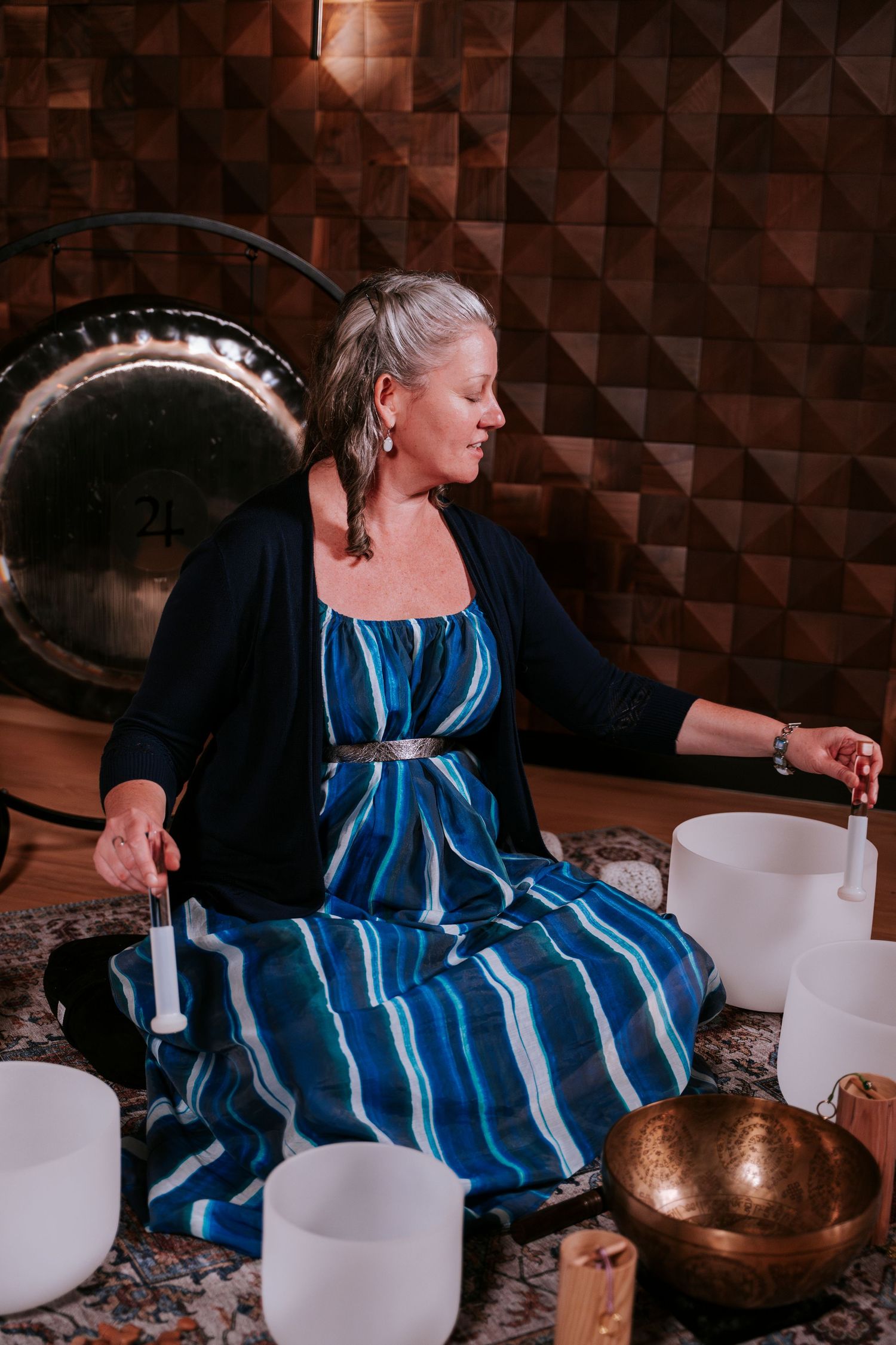 A person in a blue striped dress demonstrates traditional sound healing techniques with copper singing bowls.