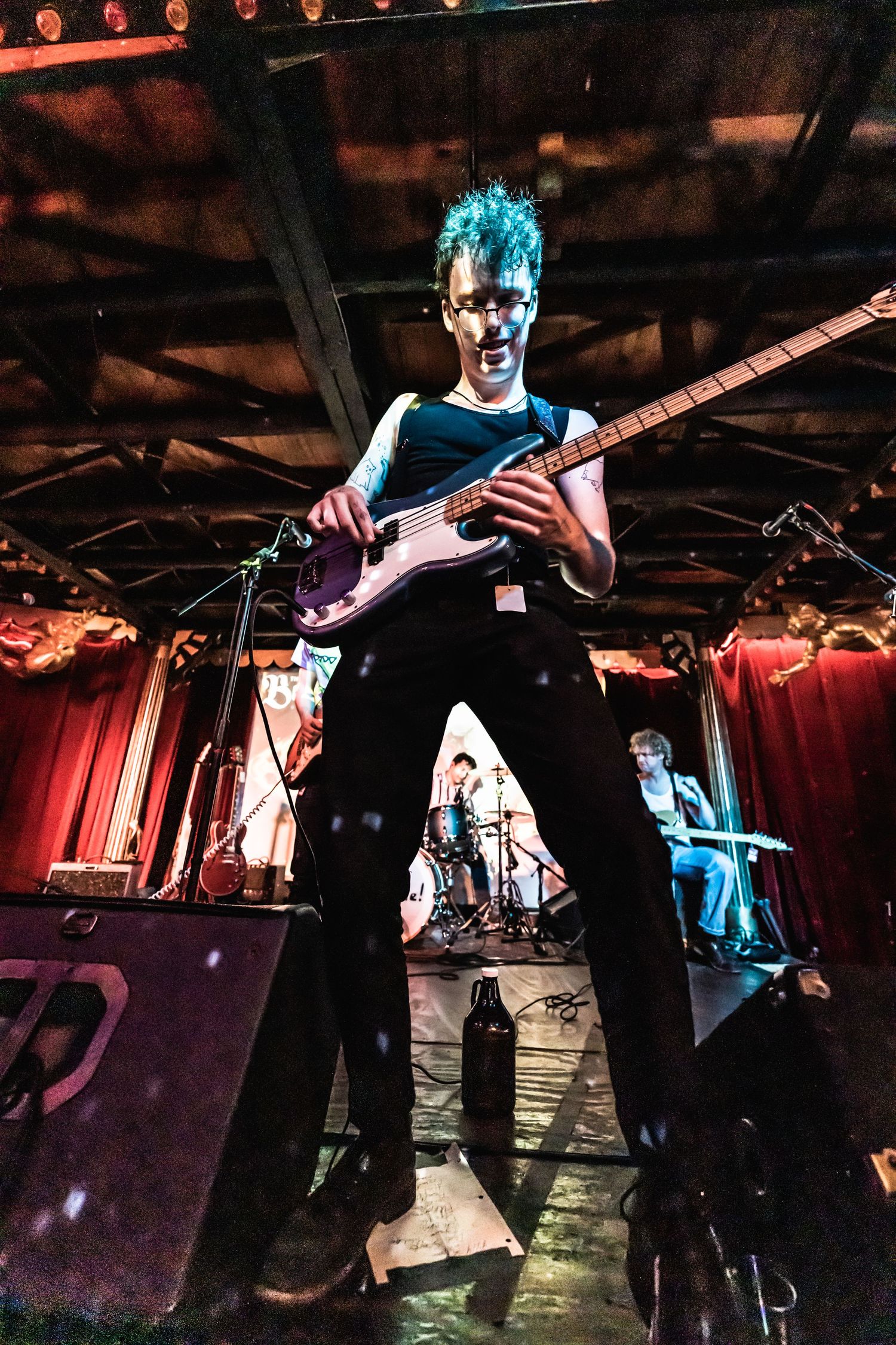 Energetic performer with bright blue hair plays bass guitar on stage in dramatic lighting.