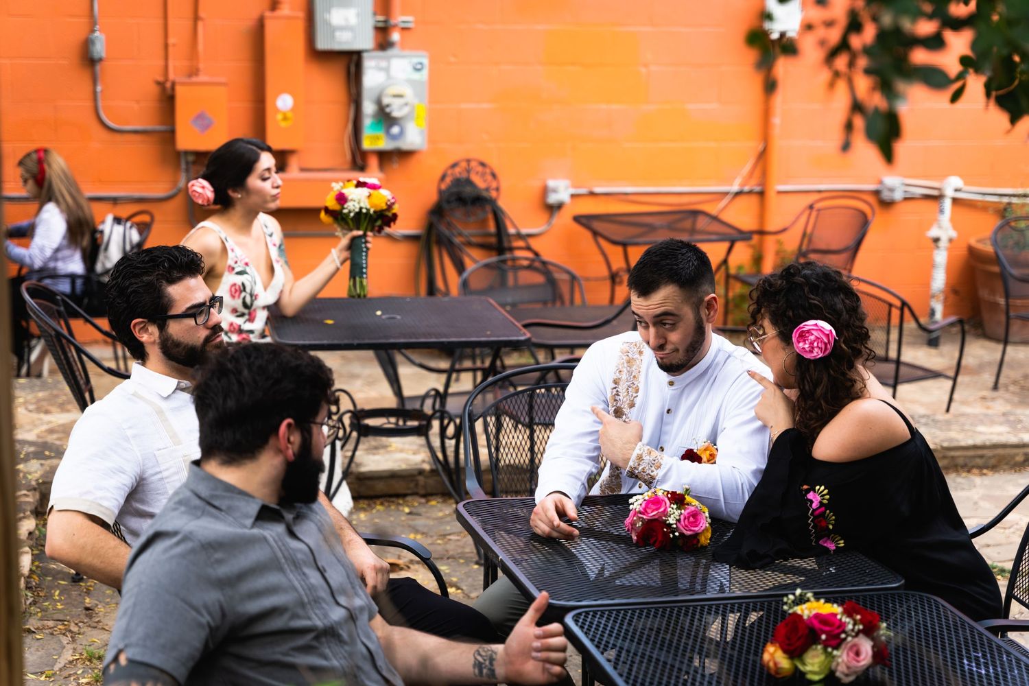 Wedding guests chatting at outdoor tables against a bright orange wall.