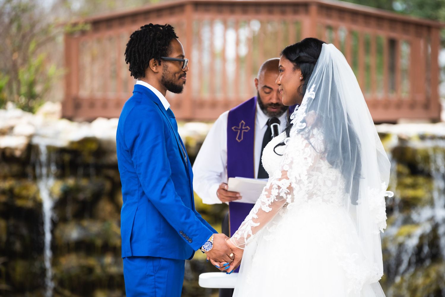 Wedding ceremony in front of waterfall backdrop with officiant in purple stole conducting service.