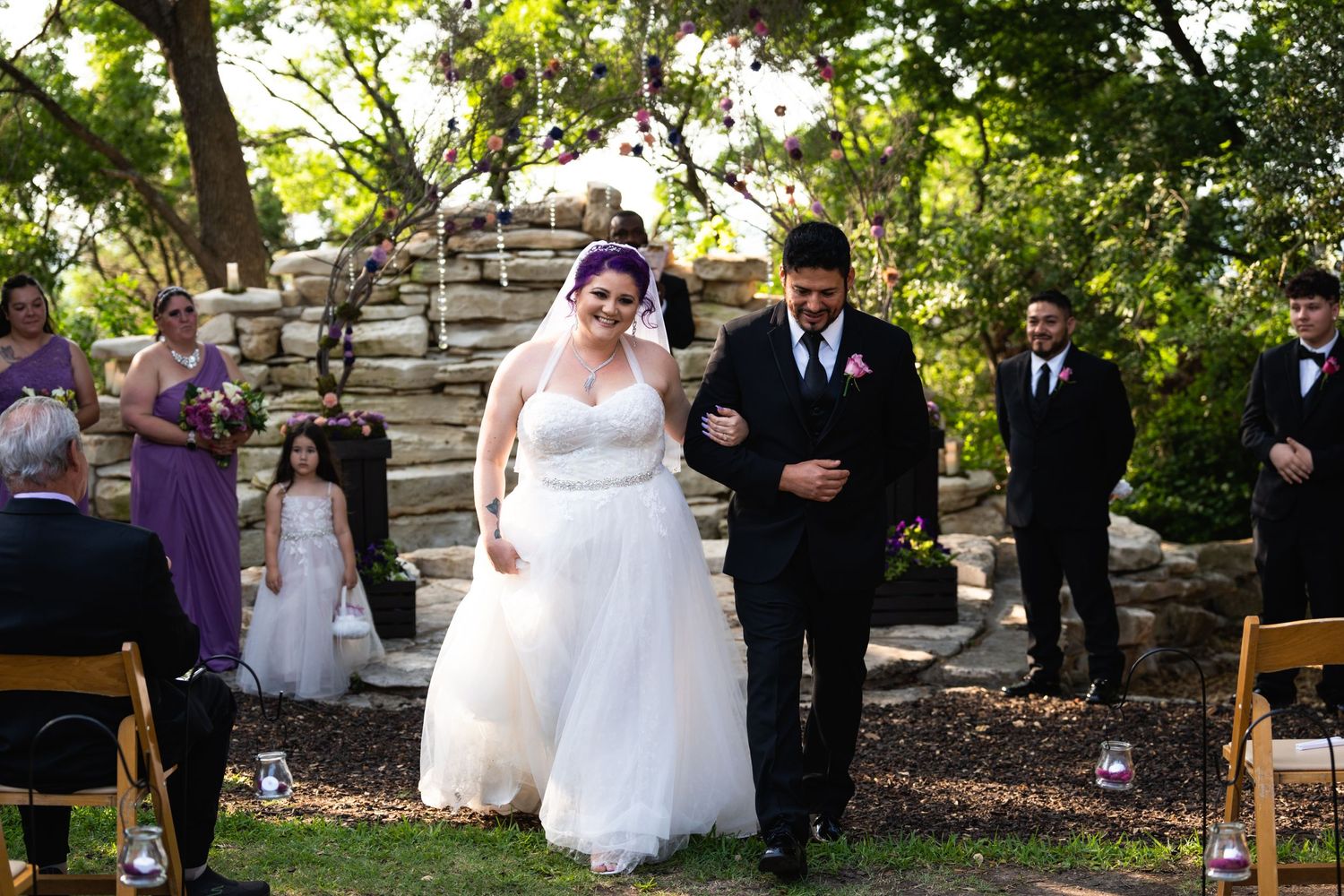 Wedding ceremony procession on stone steps with wooden chairs and natural greenery.