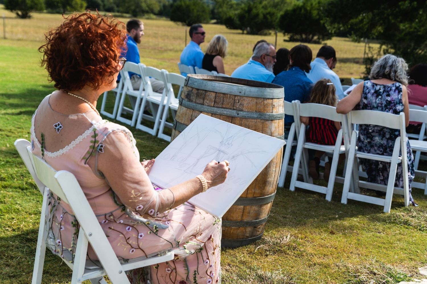 Guest sketching during an outdoor wedding ceremony with wooden chairs and rustic barrel decor.