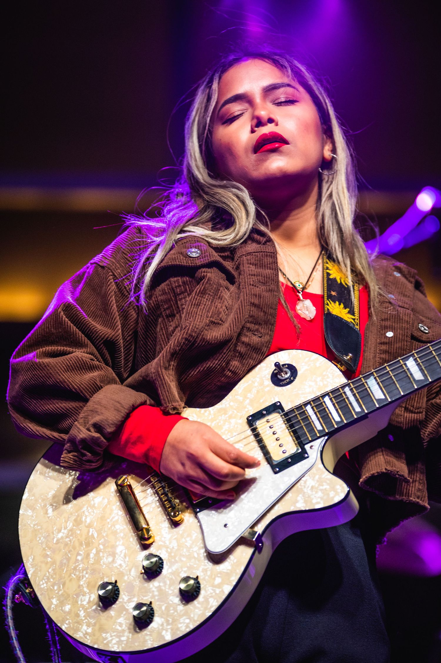 A musician performs with a white Les Paul guitar under purple stage lighting wearing a brown jacket.