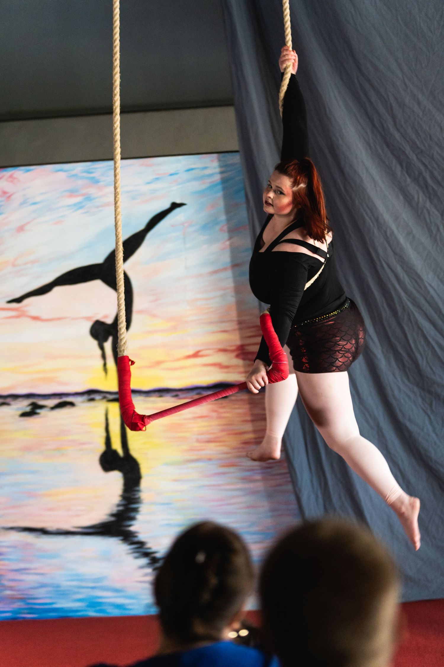 Aerial performer hangs gracefully from rope against painted sunset background.