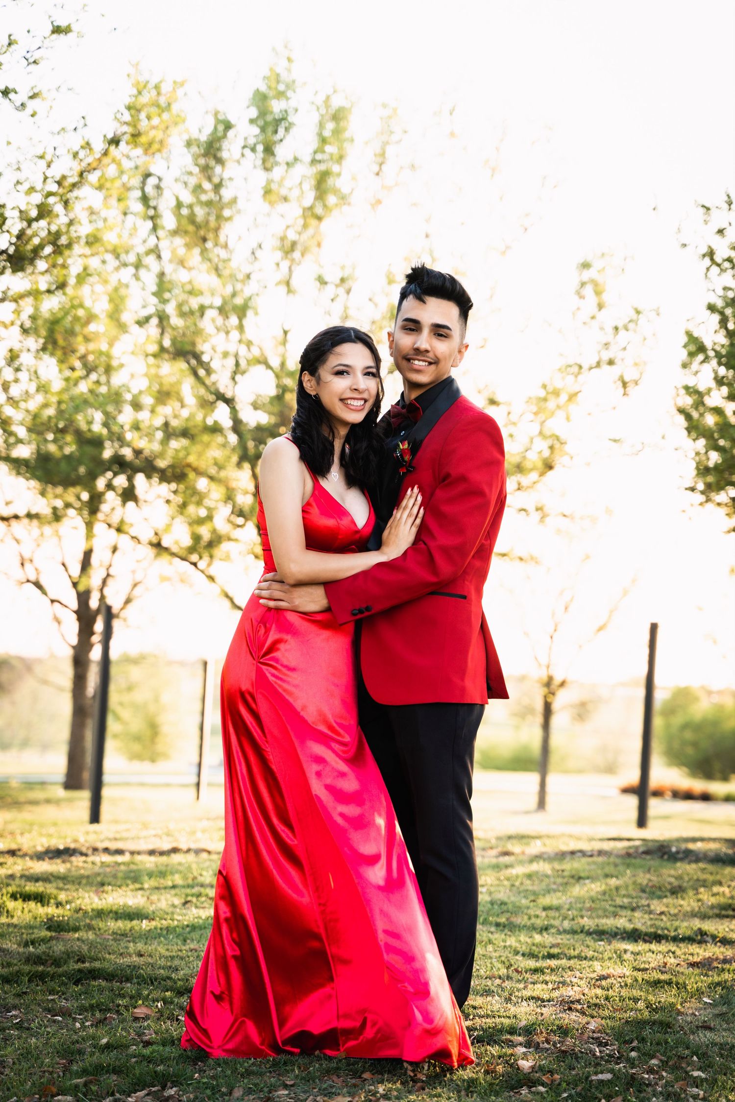 A couple in formal attire embraces outdoors, with the woman in a flowing red gown.