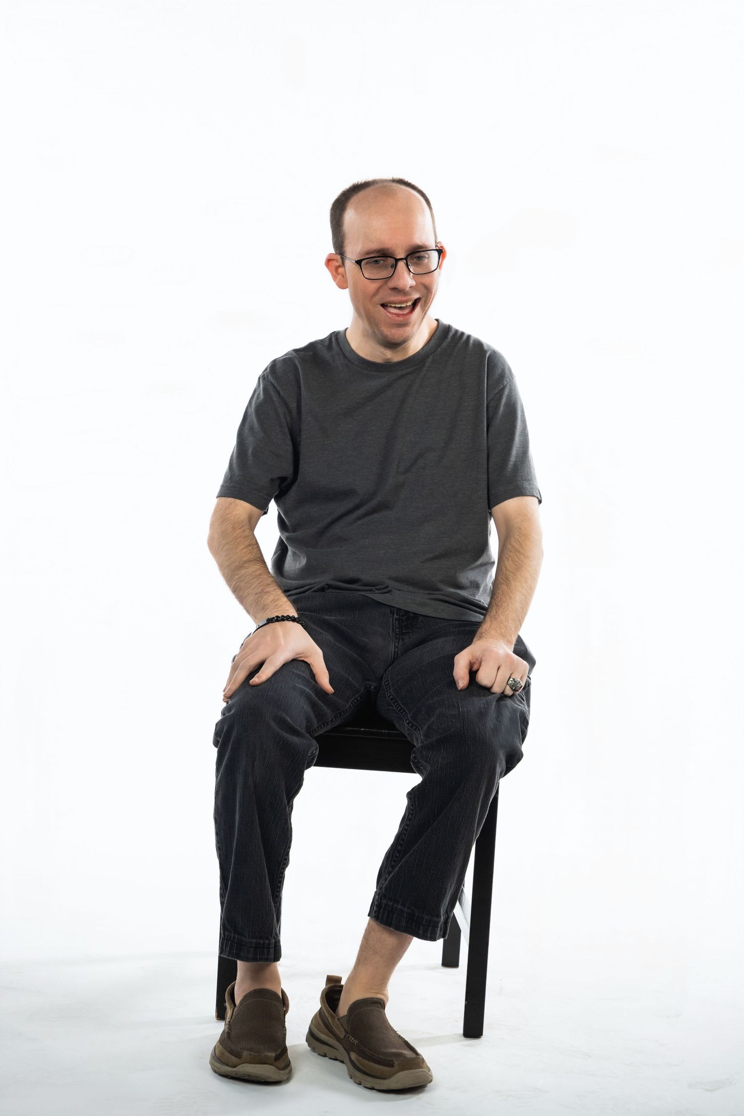 Individual sitting on a black chair wearing glasses, a grey t-shirt and cargo pants with brown shoes against white background.