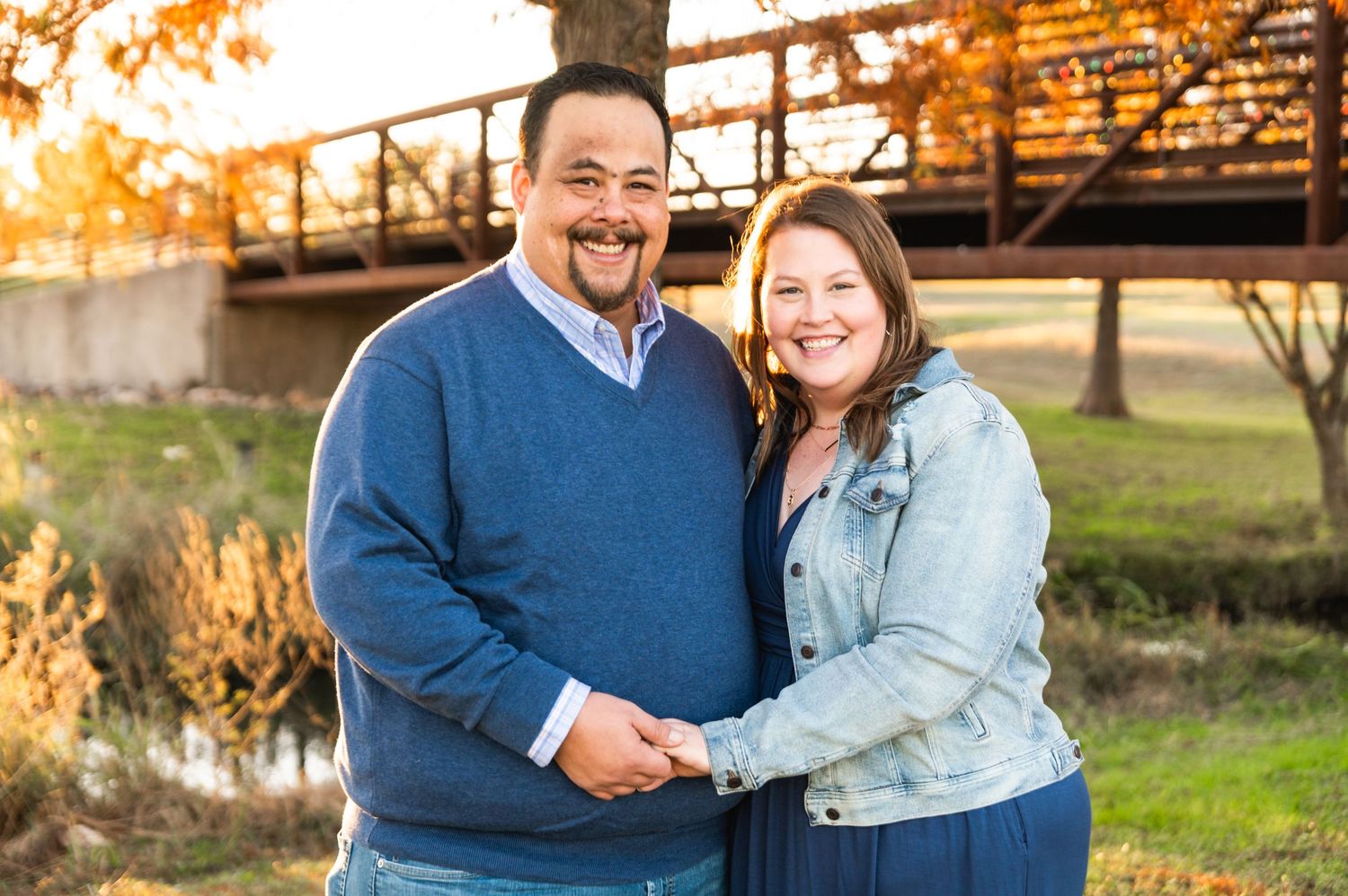 Couple in casual autumn attire poses outdoors near a bridge during golden hour.