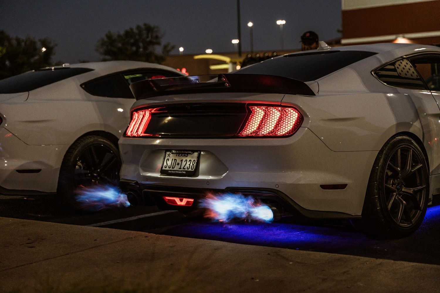 Modified Ford Mustang showing blue flame exhaust effects in dark parking lot.