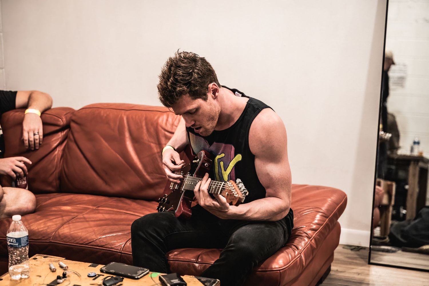 A musician plays acoustic guitar while seated on a brown leather couch.